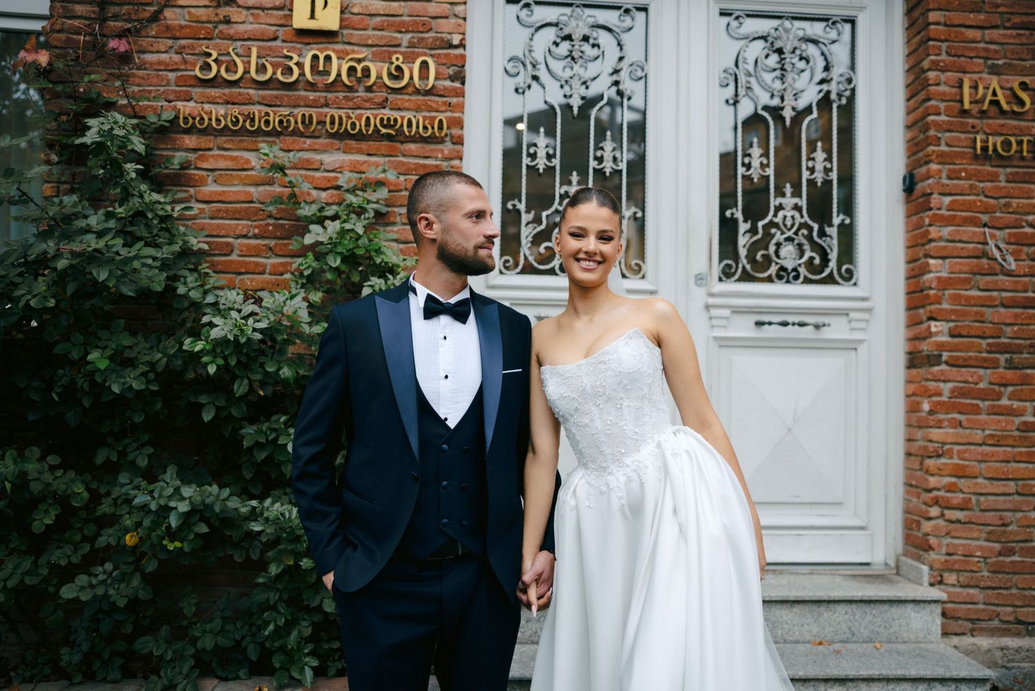 Couple poses in front of ornate brick building with decorative iron door.