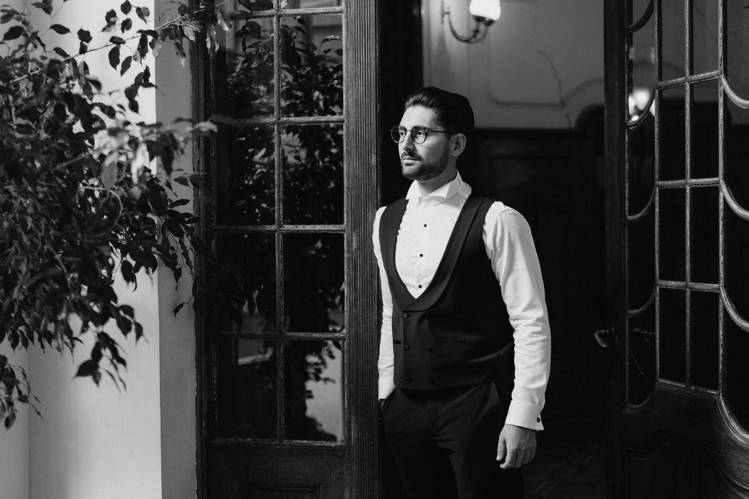 Man in formal black and white attire standing in front of a window in dramatic lighting.
