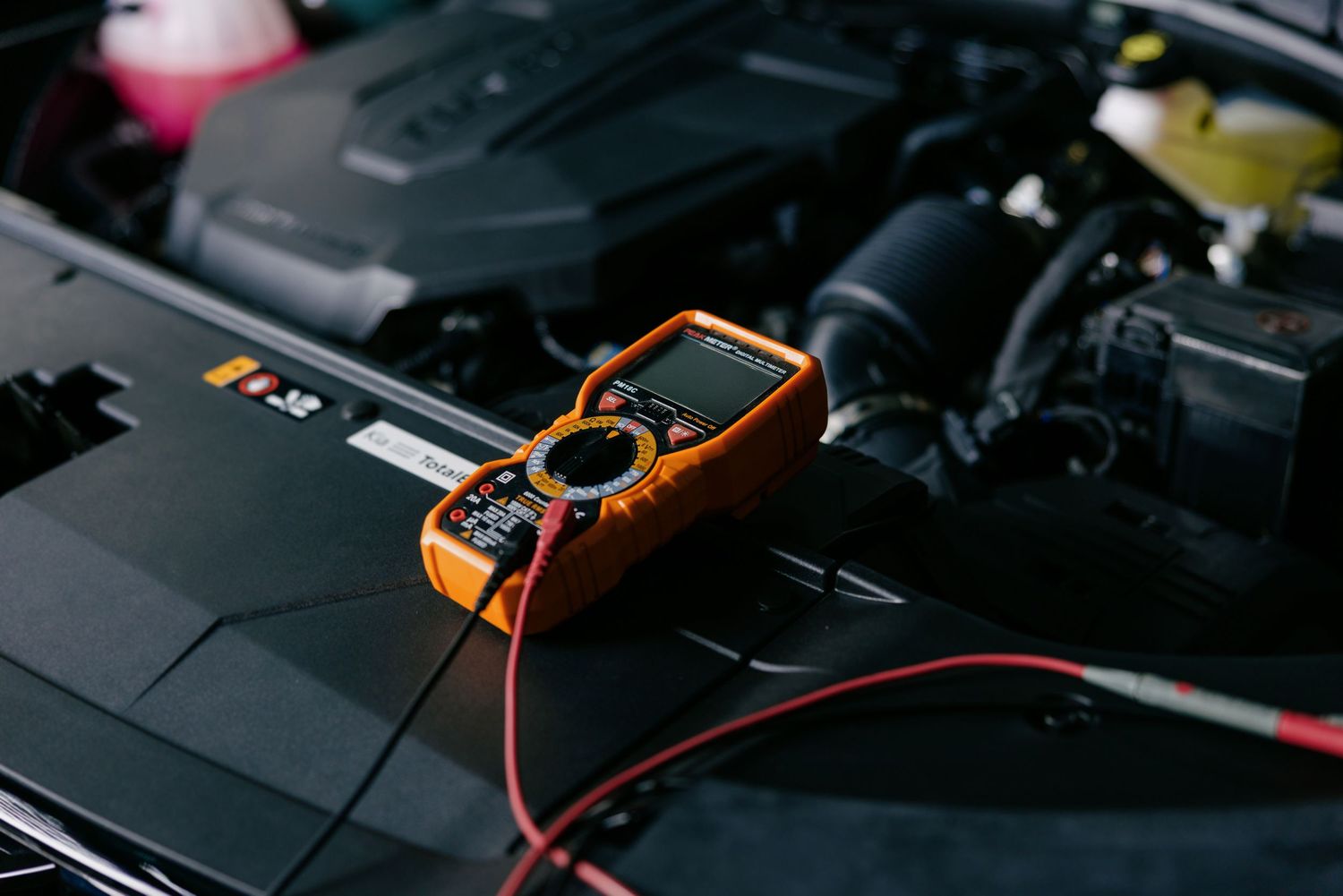 Orange multimeter with red probes testing a car battery in an open vehicle engine bay.