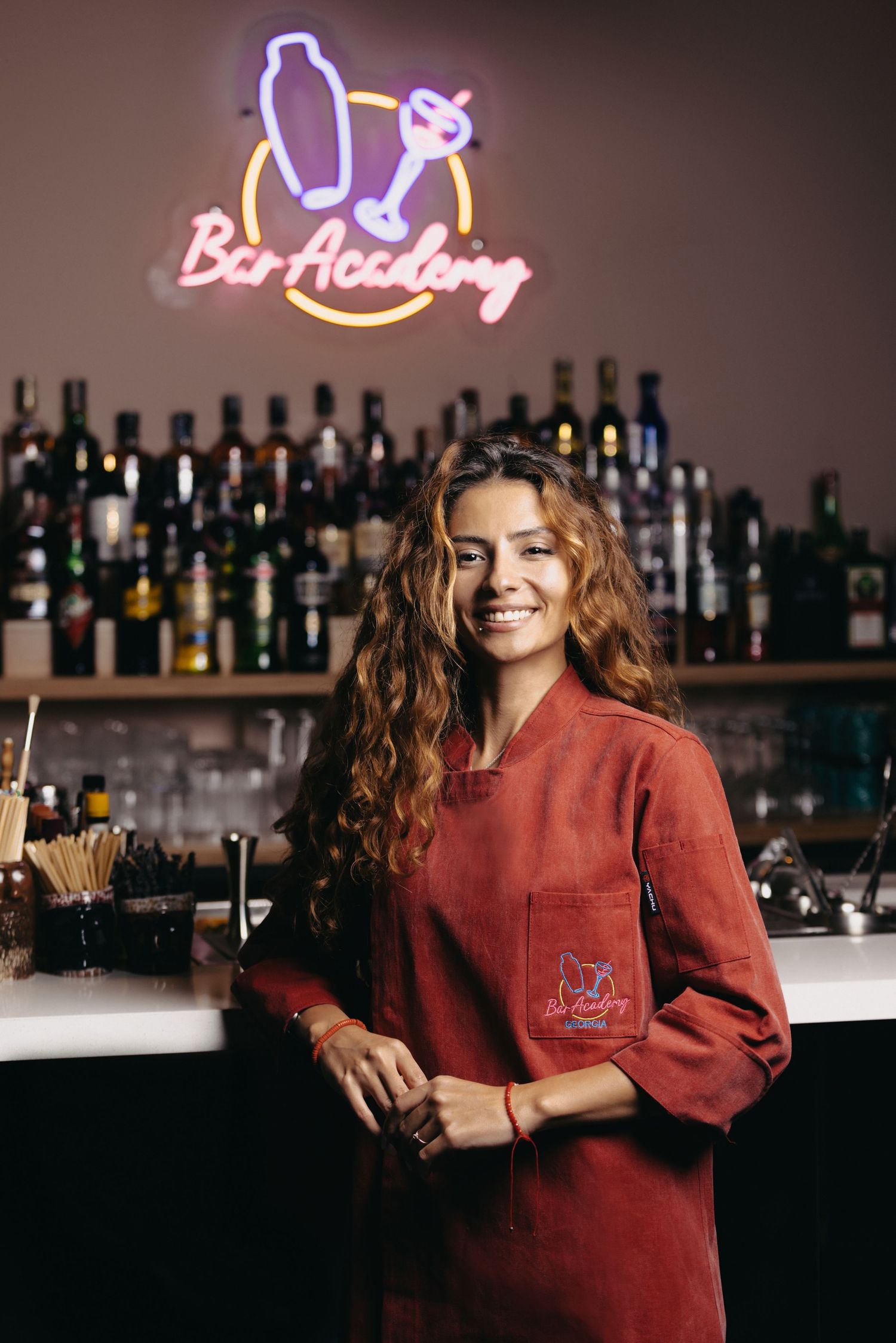 Bar staff member in red uniform smiling while working at cocktail counter.