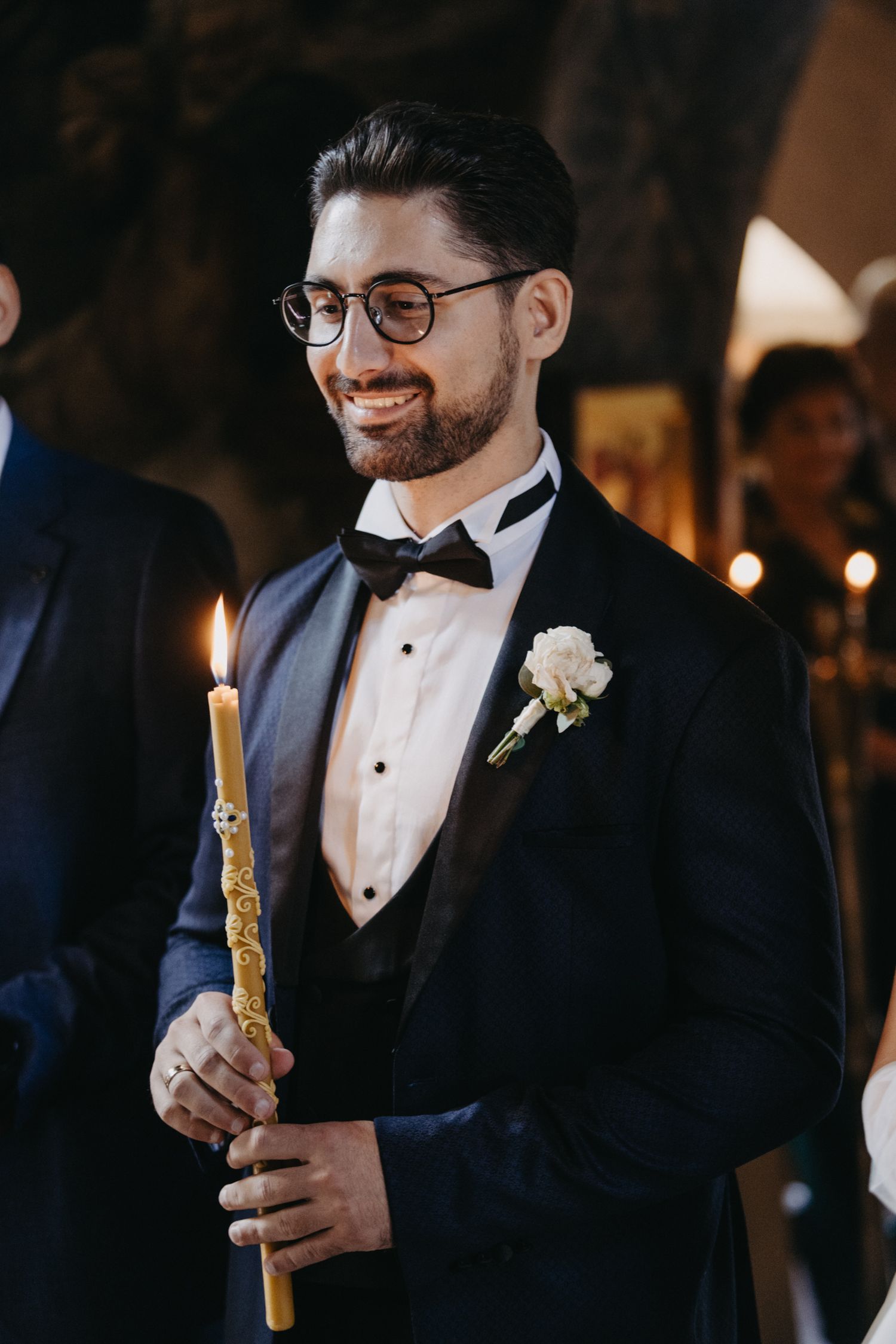 Groomsman in tuxedo and glasses holds decorated candle during traditional ceremony.