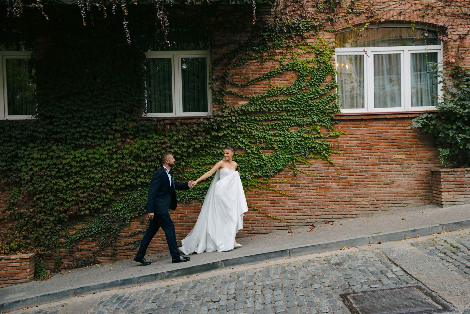 Newlyweds walking together along an ivy-covered brick building wall.