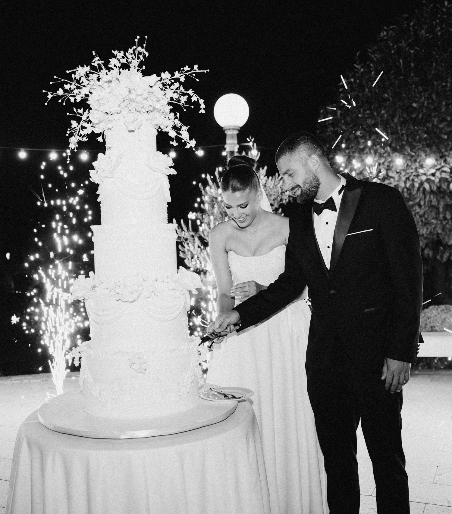 Wedding couple cutting their elegant white tiered cake with sparklers in the background.