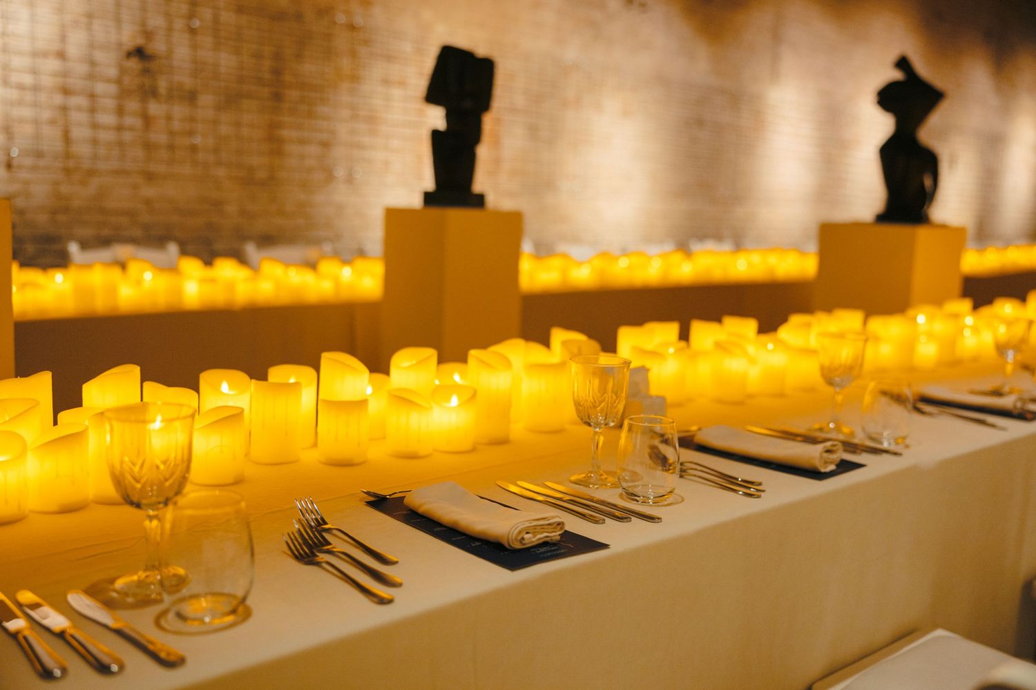 Elegant table settings with rows of illuminated candles in a brick-walled dining space.
