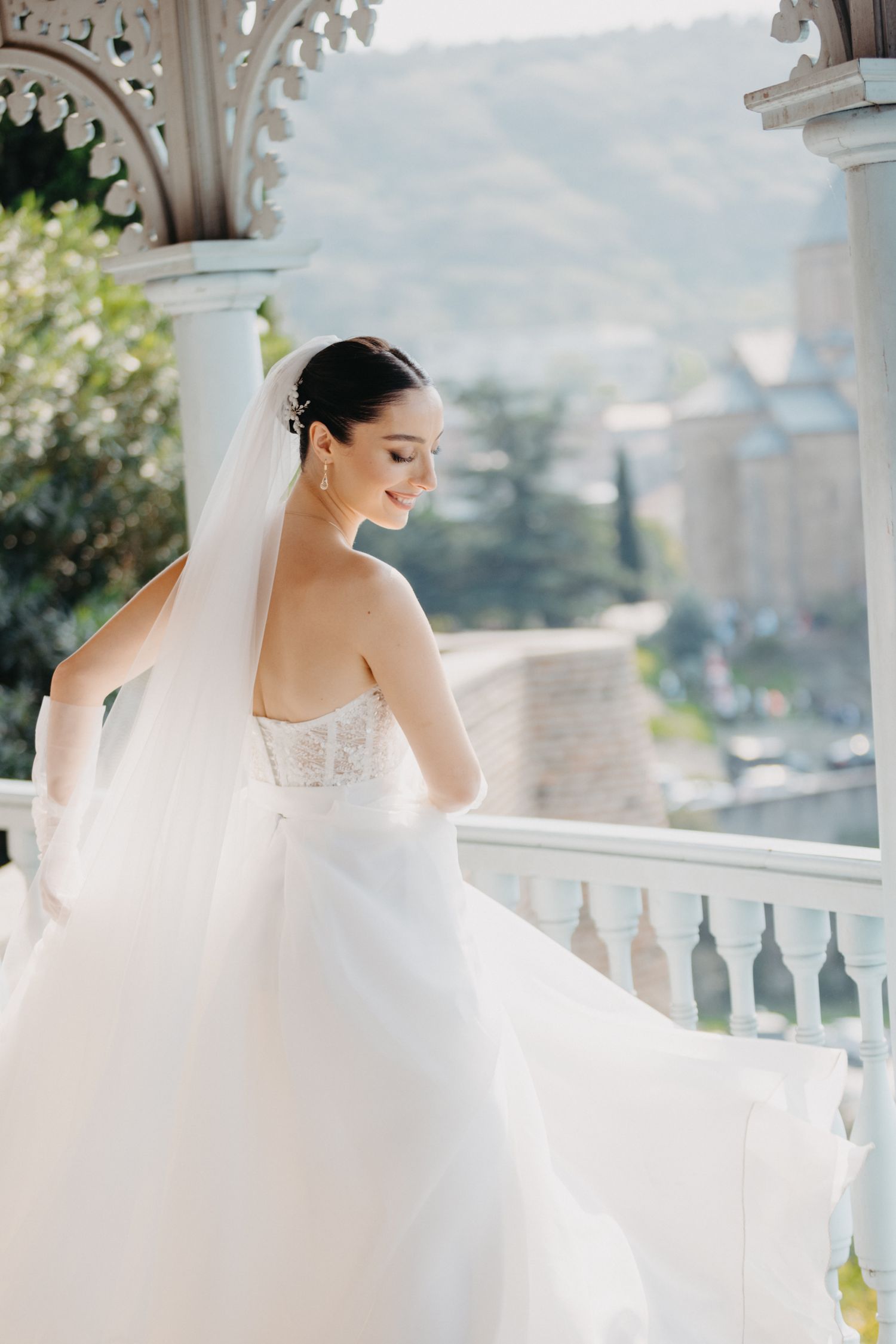 Elegant bride in a strapless white gown poses on a balcony with ornate architecture and scenic mountain views.