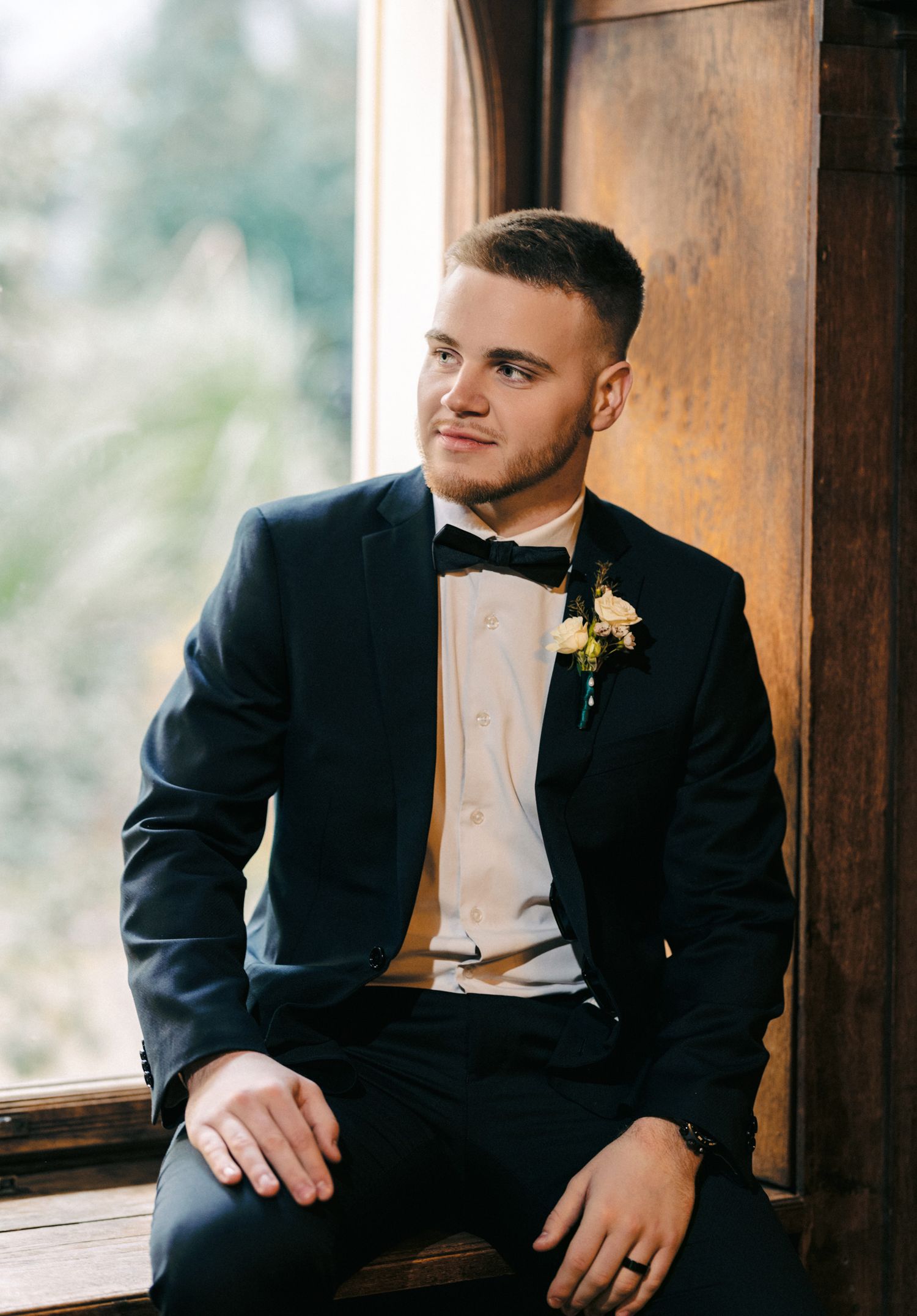 Stylish groom in navy tuxedo sits by window with boutonniere.