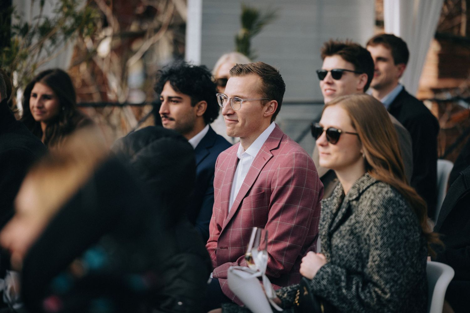 Wedding guests in formal attire seated outdoors during daytime ceremony.