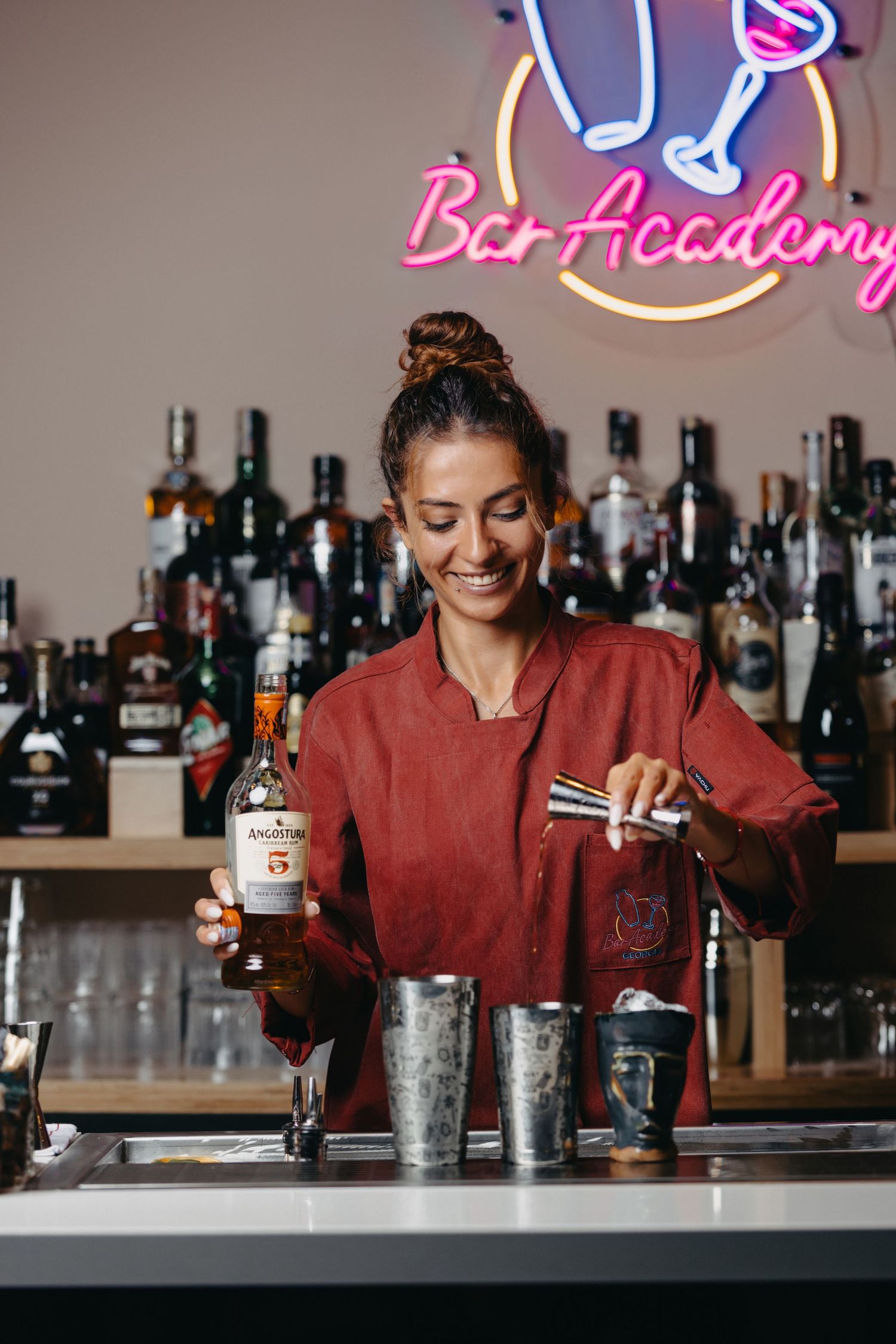 Bartender in red uniform mixing drinks at bar counter with neon signs in background.