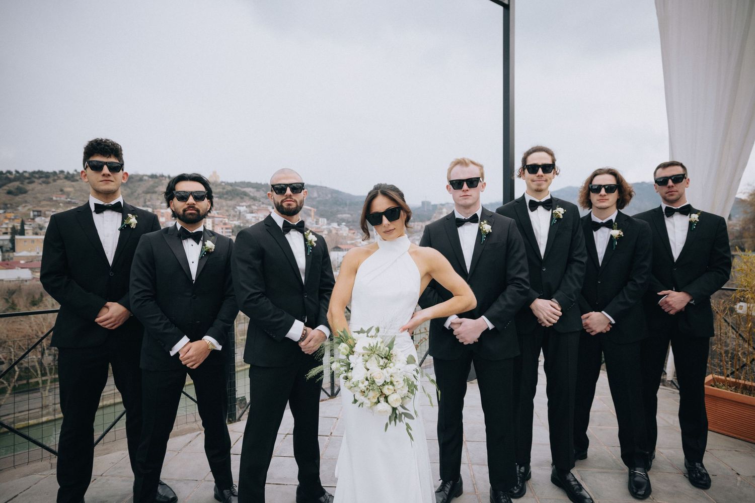 Wedding party in formal black attire poses with bride on mountain overlook terrace.