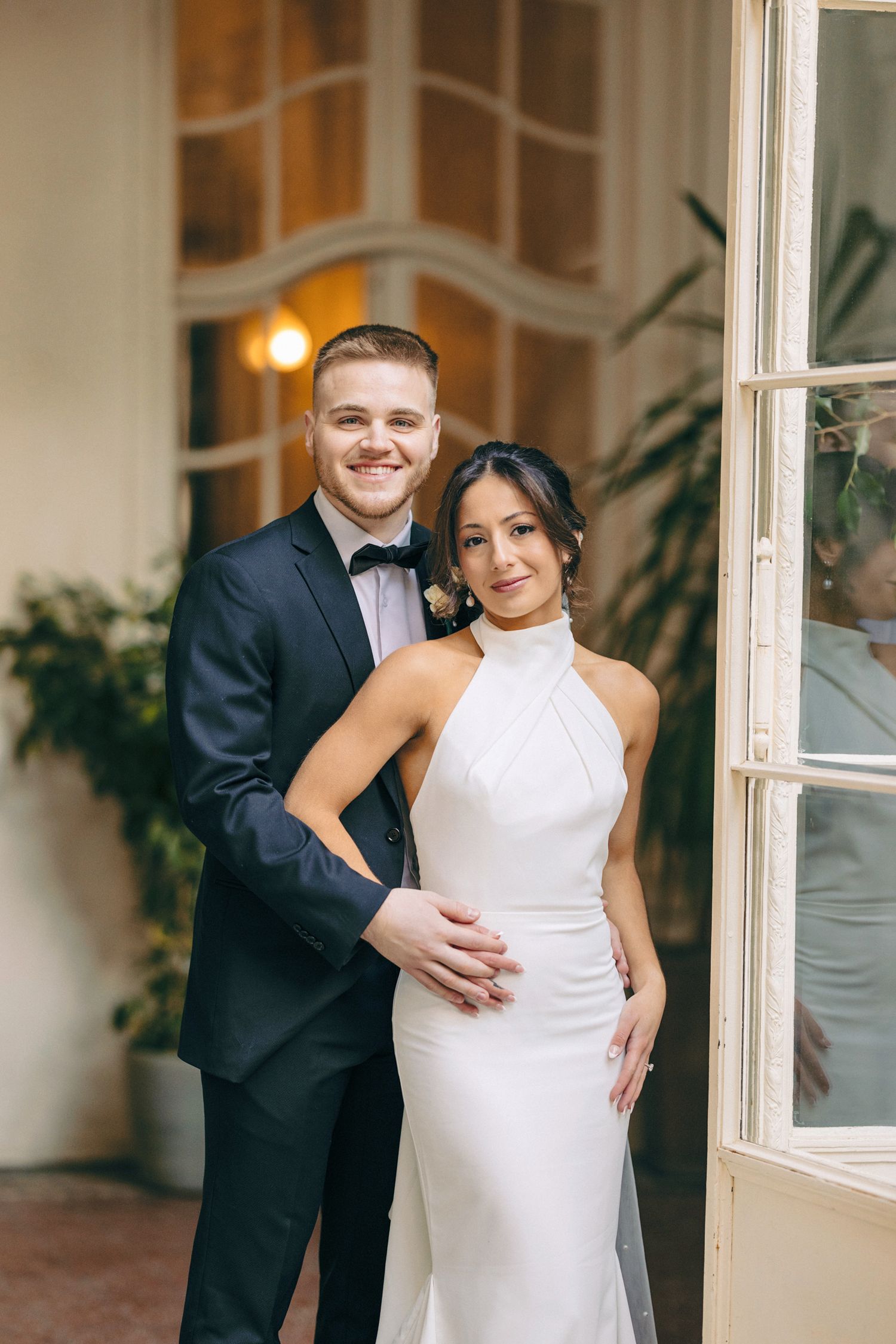 Wedding couple embraces in bright interior with white walls and windows.