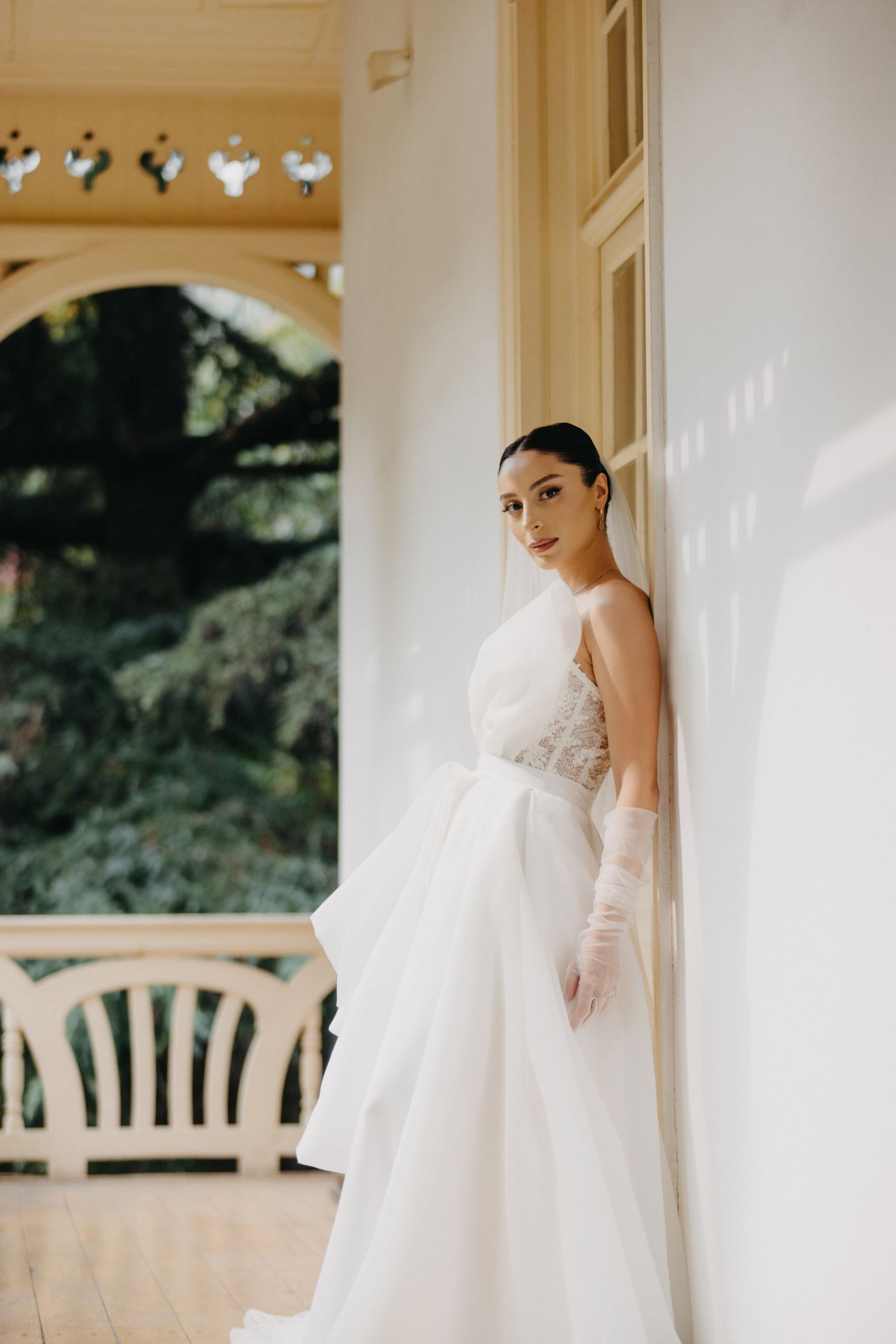 Bride in flowing white gown poses elegantly along white columned porch with natural lighting.