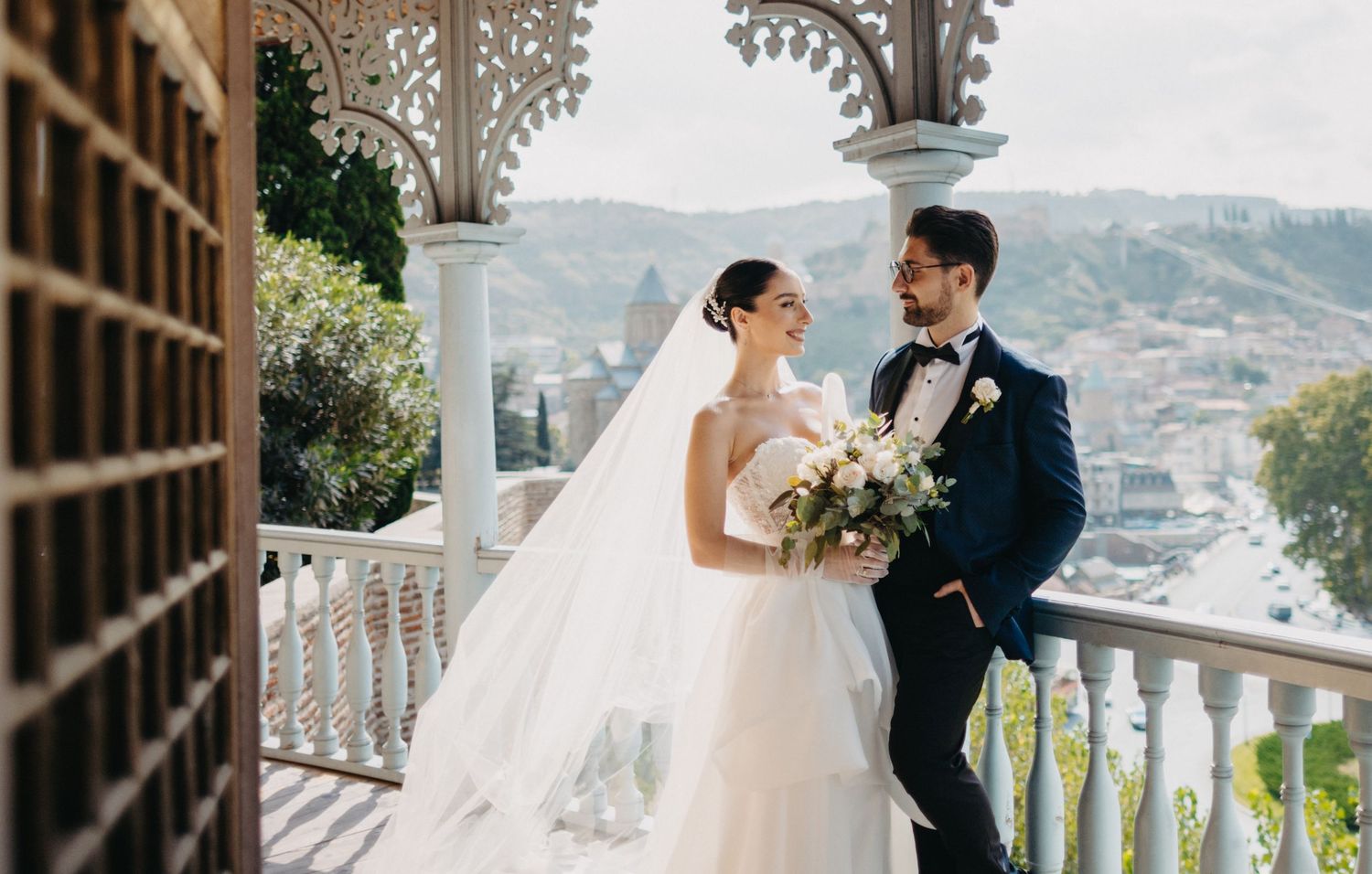 Wedding couple embraces on an ornate balcony overlooking a scenic mountain landscape.