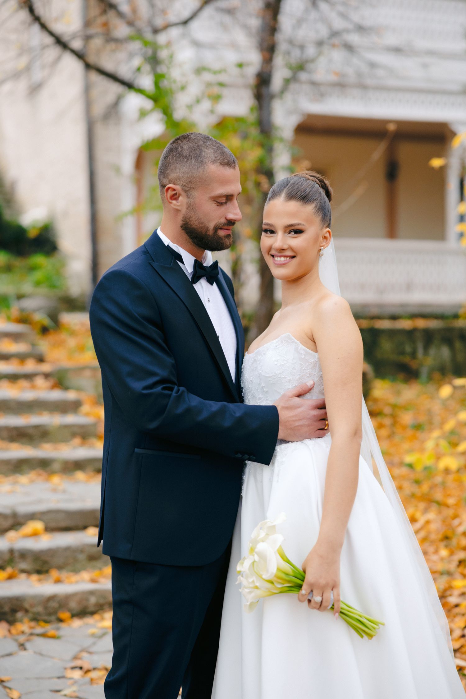 A couple in wedding attire embraces on stone steps surrounded by autumn foliage.