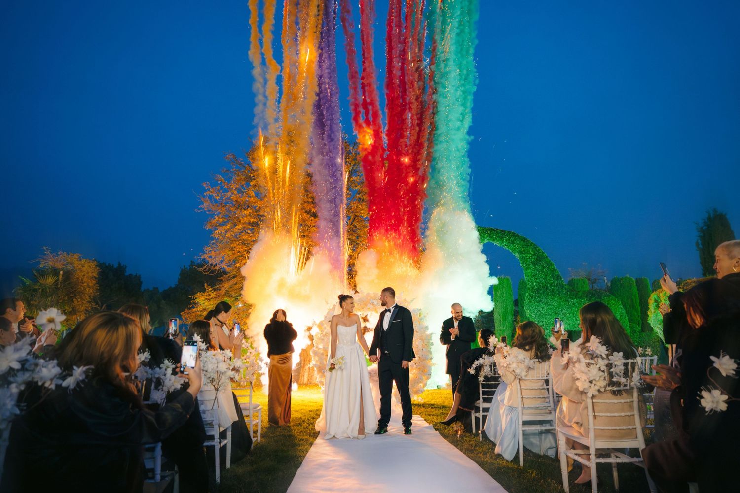 Colorful smoke bombs create vibrant streaks against a blue sky at an outdoor wedding celebration.