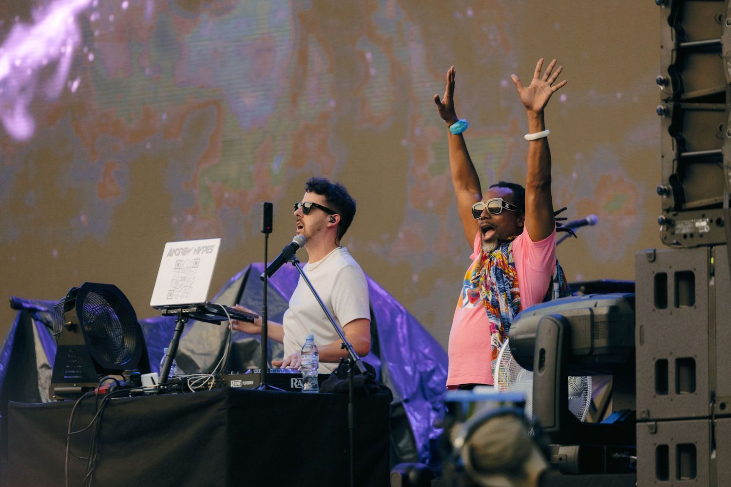 DJs performing on stage with raised hands and electronic equipment at a music festival.
