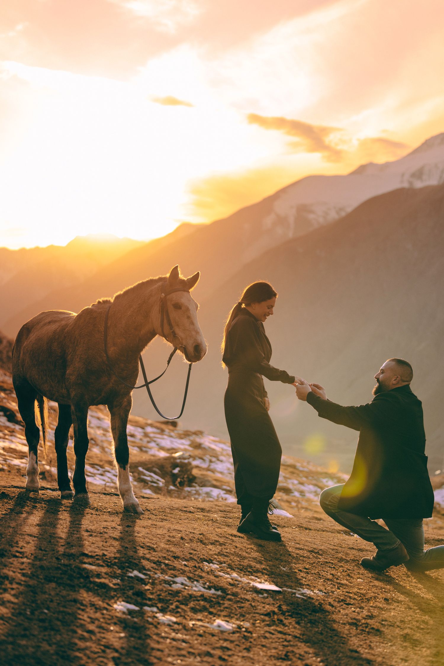 Two people kneel in the snow at sunset with horses silhouetted against mountain peaks.
