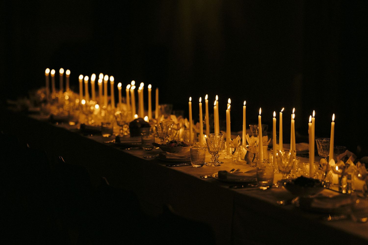 Multiple lit candles create a warm ambiance in a dark room at a formal dinner setting.