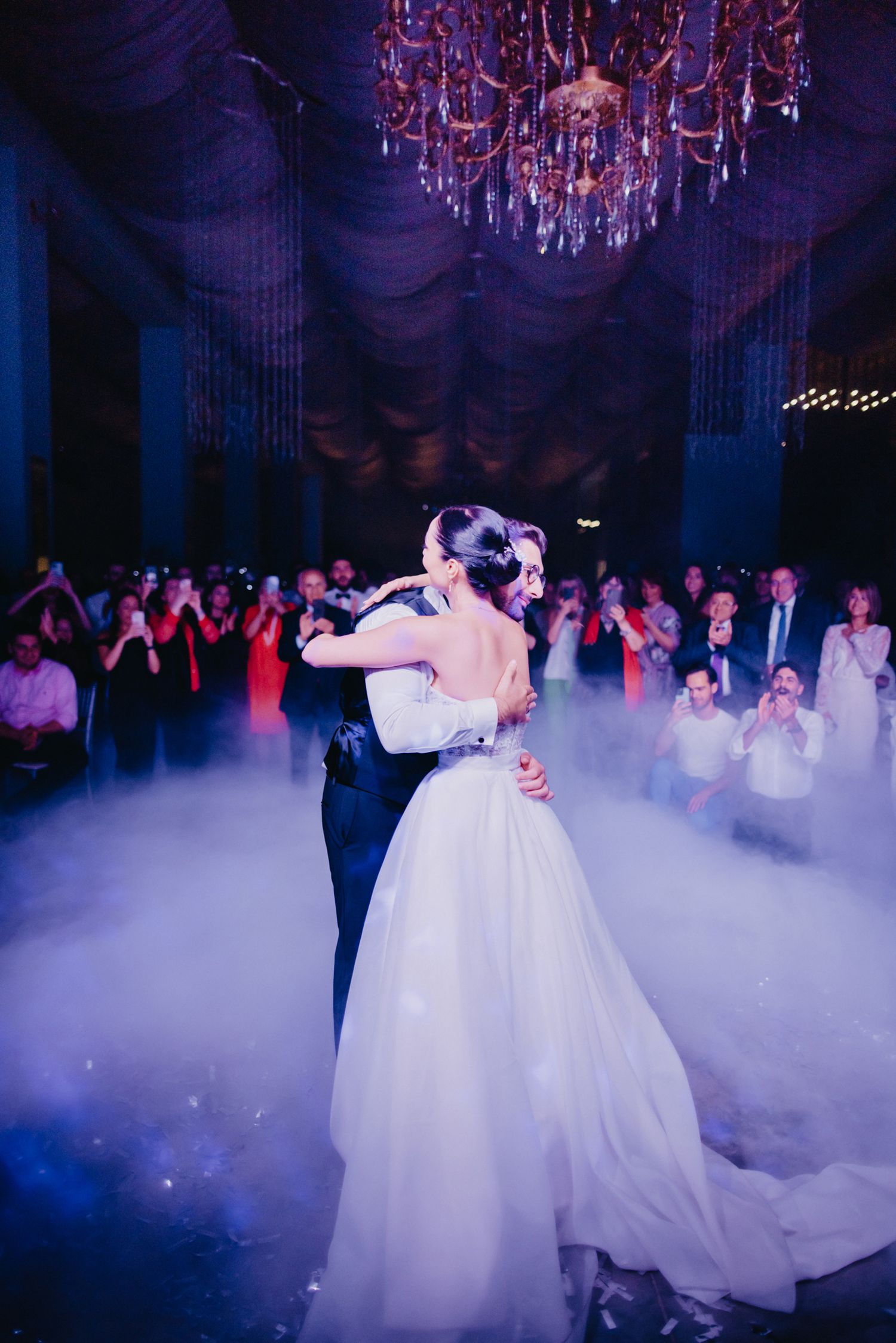 First dance of newlyweds under crystal chandelier with dramatic fog effect.
