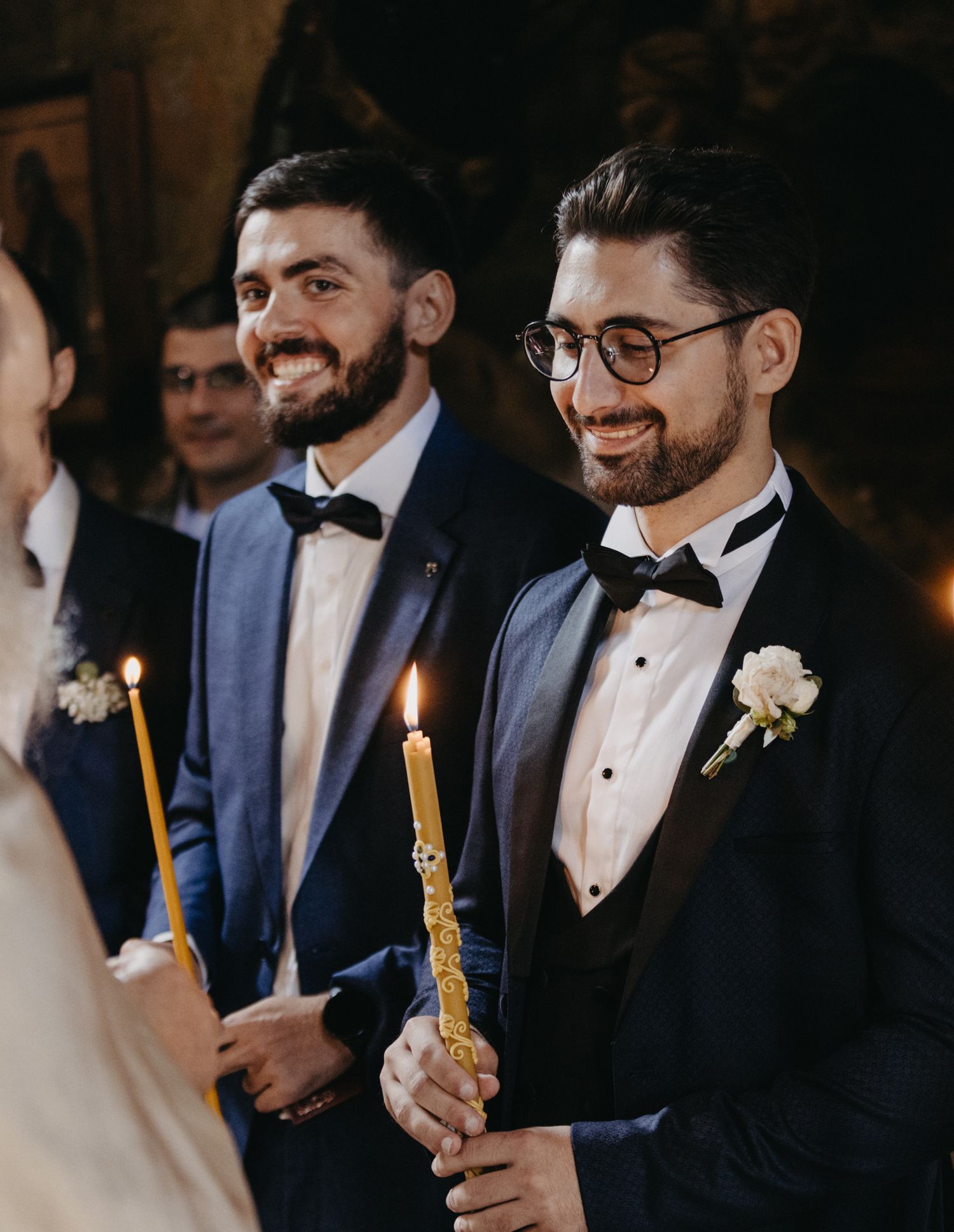Groomsmen in dark suits and bowties hold lit candles during wedding ceremony.