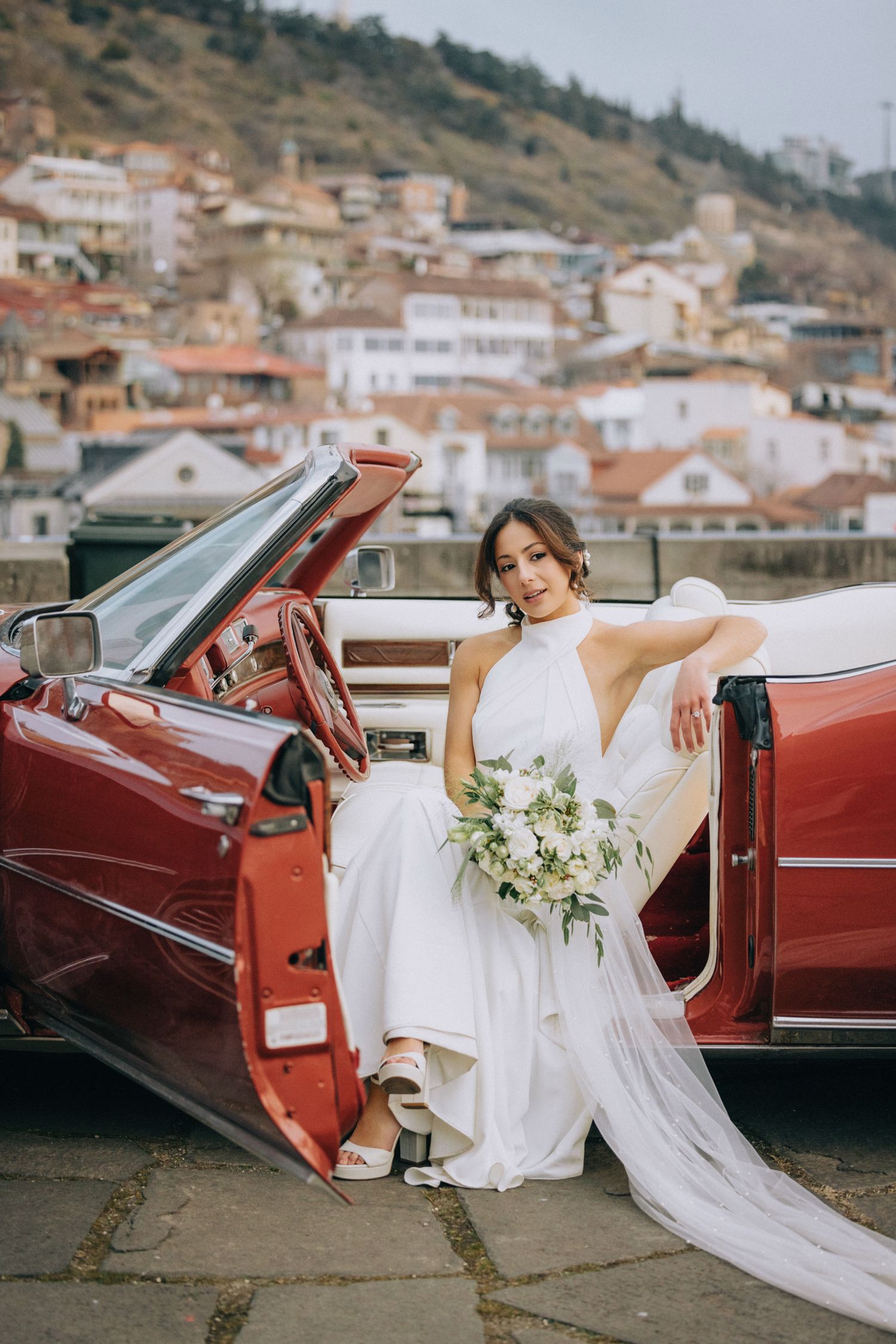 Flowing white wedding gown cascading over the side of a vintage red car with hillside buildings in background.