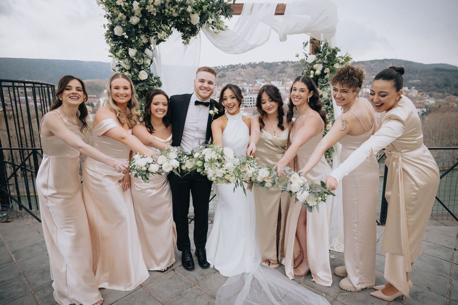 Bridal party in champagne dresses gathers around couple for group photo on scenic terrace.