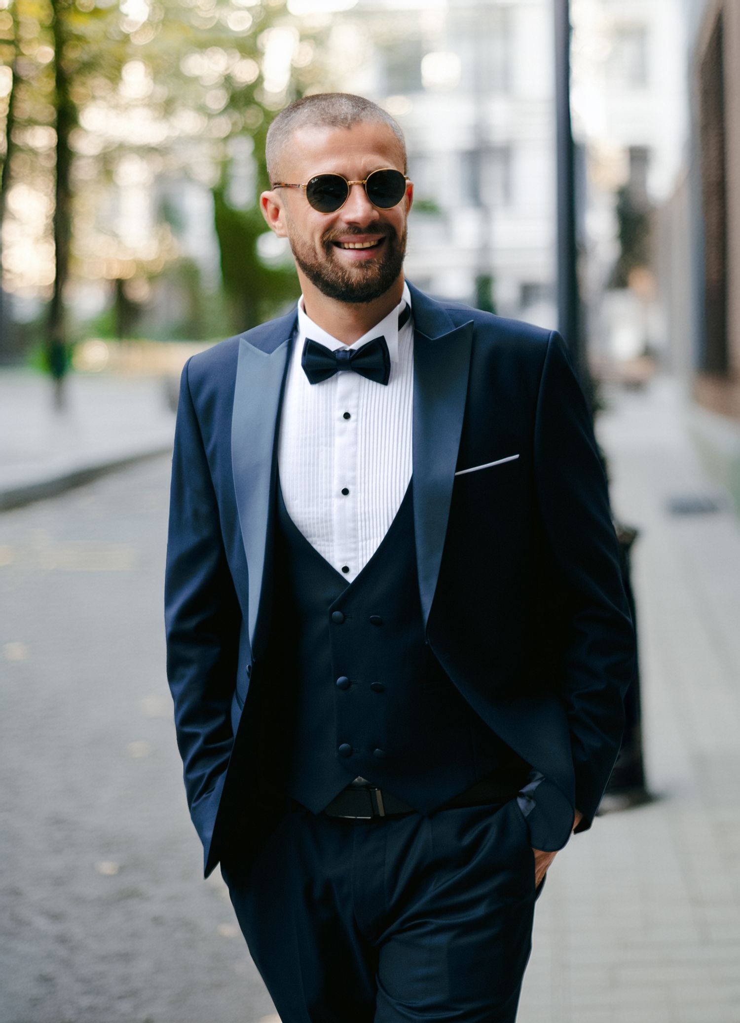 Groom in formal black tuxedo and sunglasses poses outdoors on a city street.