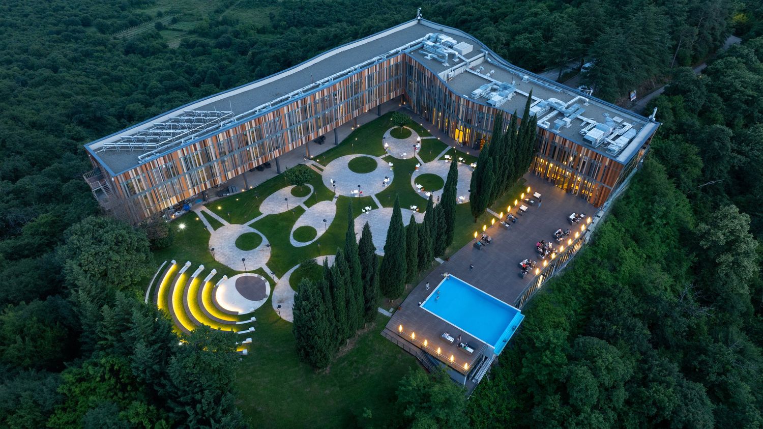 Drone shot of an illuminated resort property surrounded by lush forest and featuring a swimming pool.