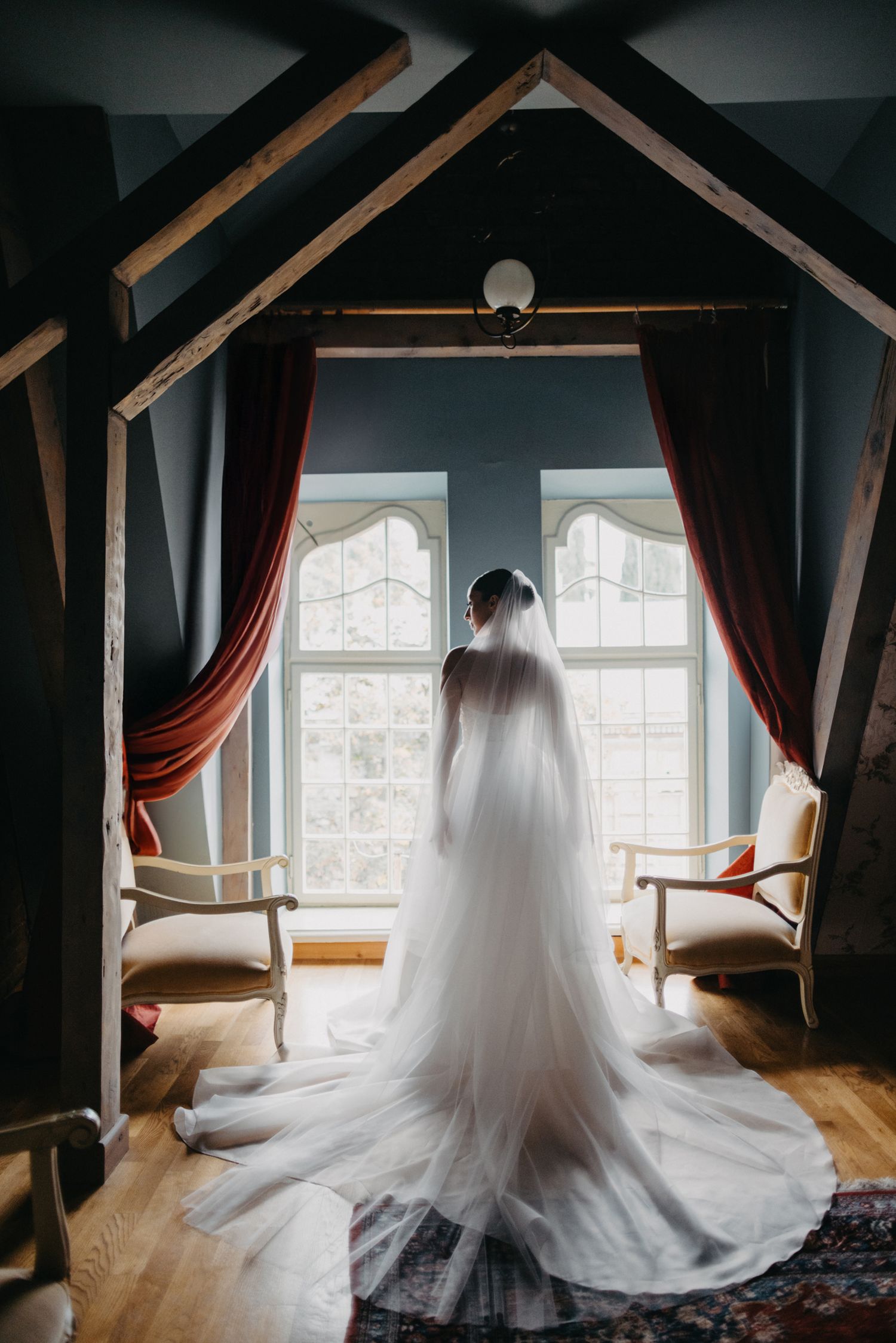 Bride in flowing wedding gown and veil standing before windows framed by red curtains and wooden beams.