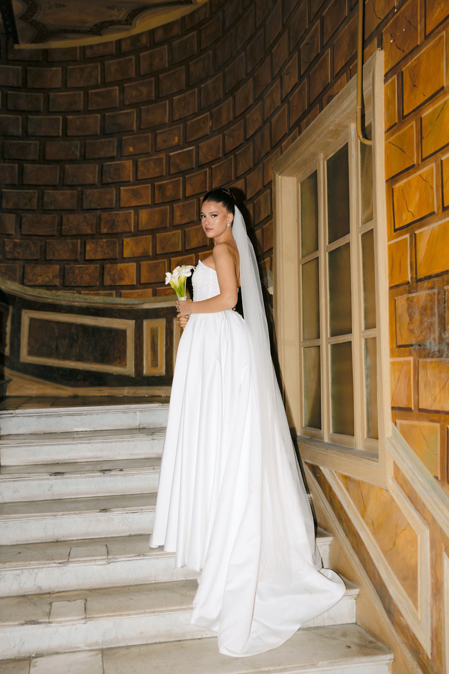 Elegant bride in flowing white dress with long veil stands on curved staircase.