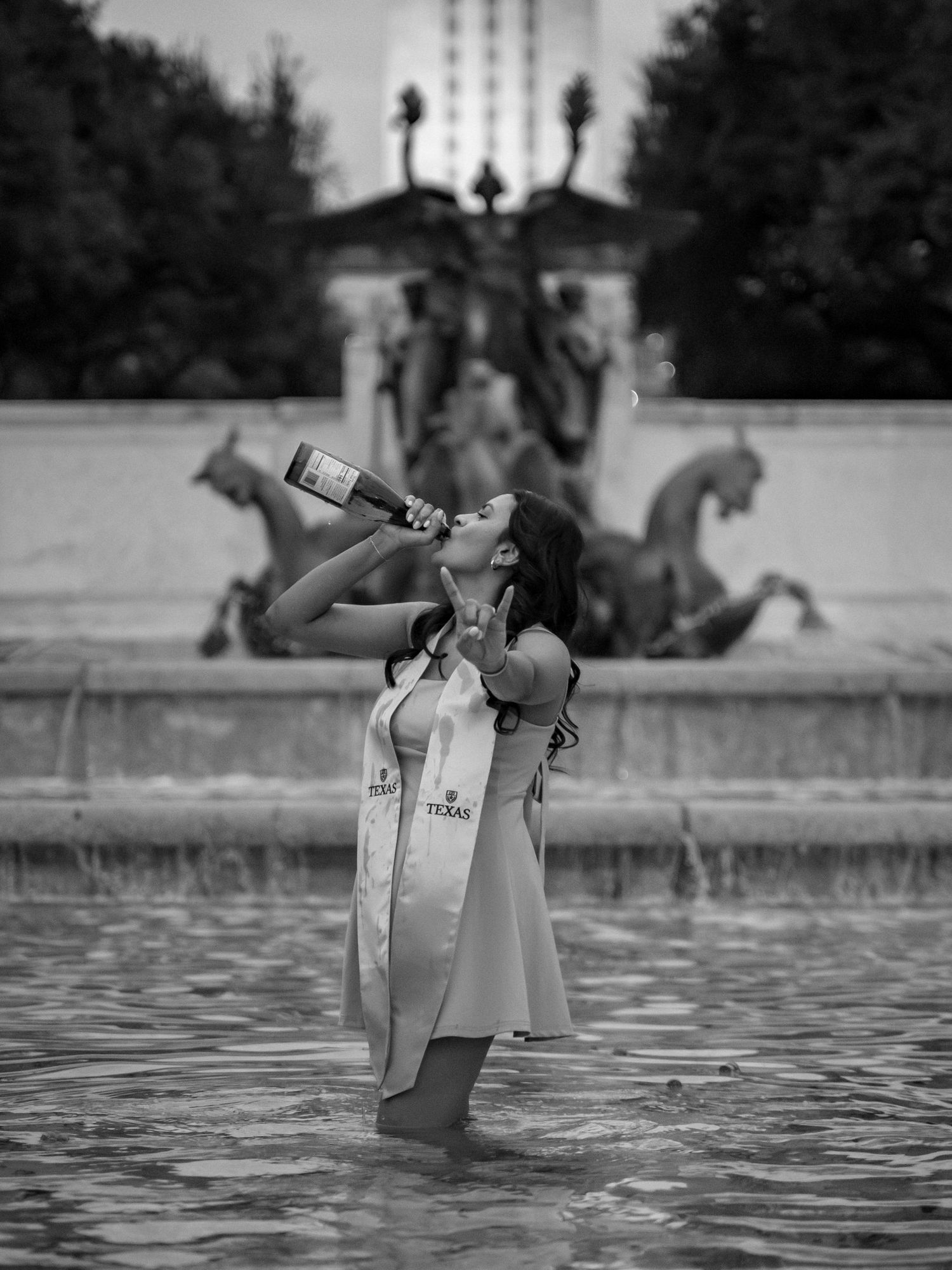 A figure in a white dress stands in a fountain while drinking from a bottle in a black and white artistic photograph.