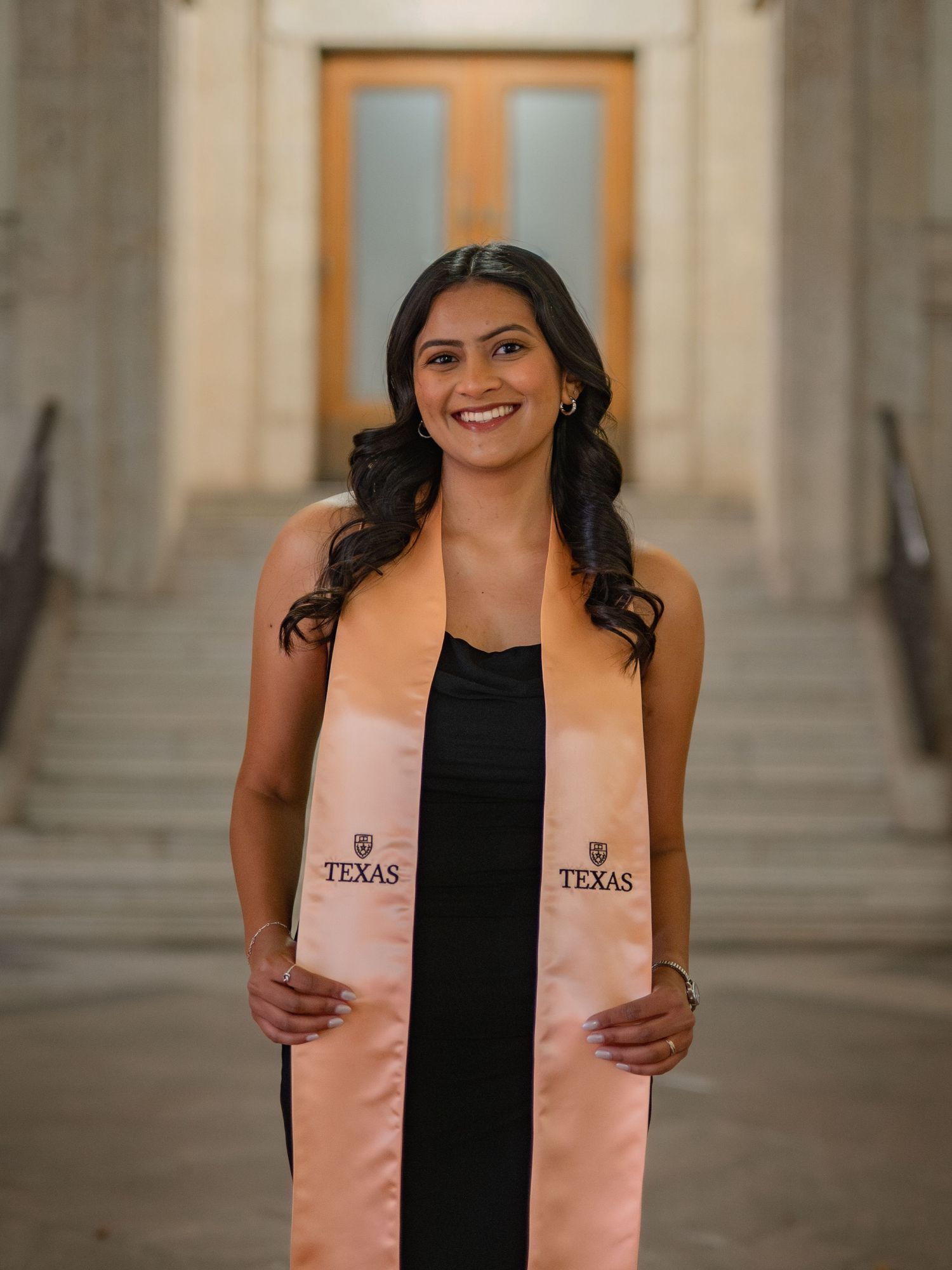 Graduate wearing a black dress and peach-colored graduation stole stands in front of stone columns at University of Texas.