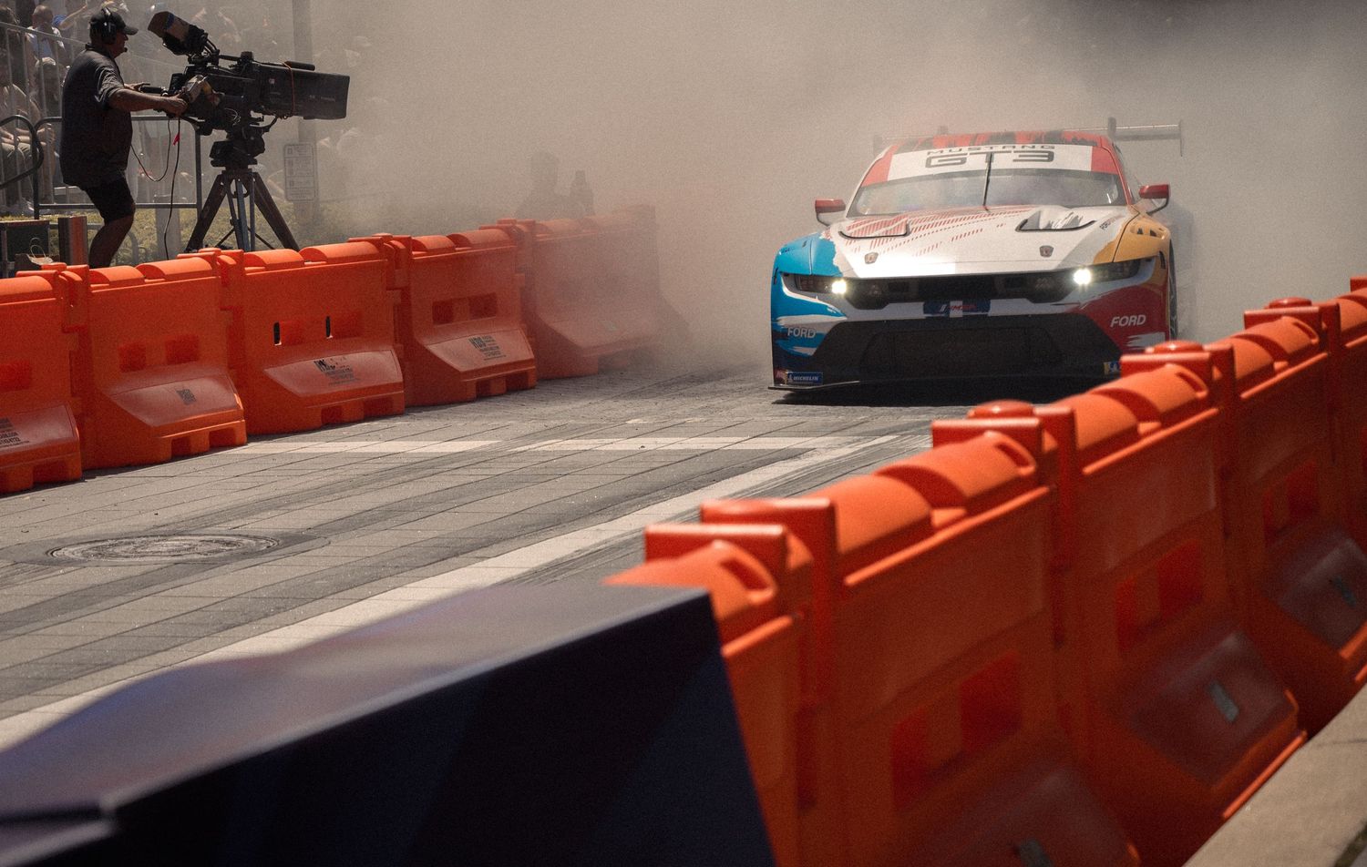 Urban street track with orange safety barriers and spectators lined up along the course.