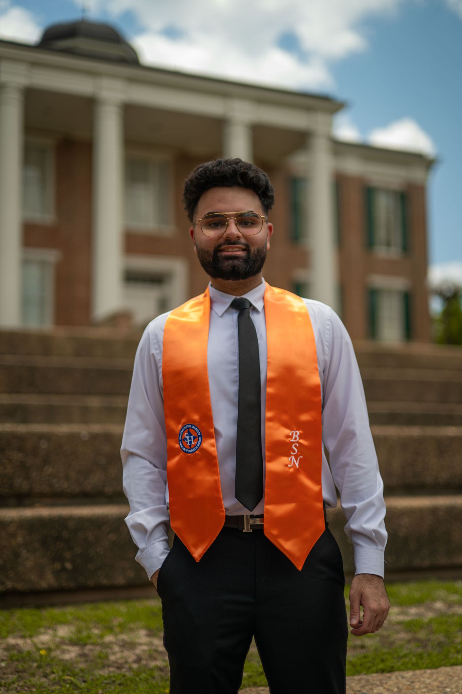 Graduate wearing orange academic stole stands proudly before brick university building.