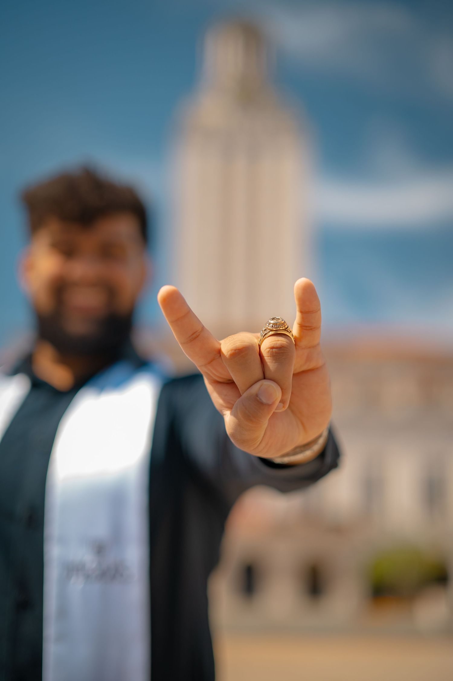 Close-up of hand making hook 'em horns gesture against blue sky and tower backdrop.
