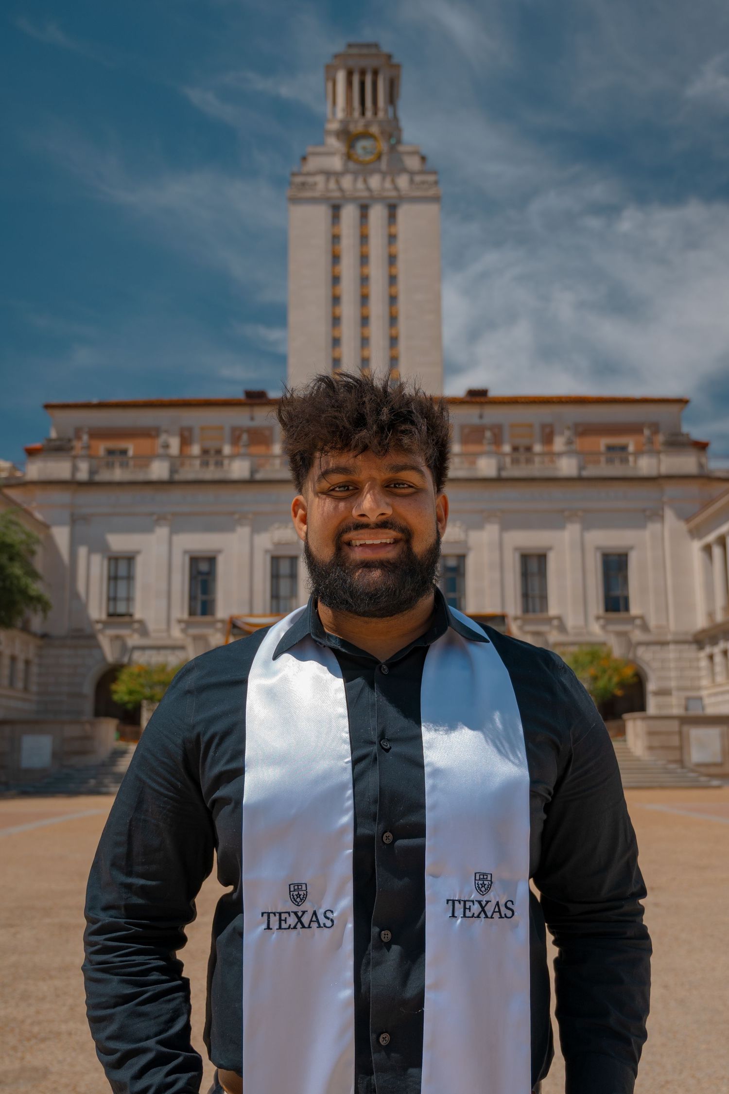 Graduate wearing Texas sash and black jacket smiles in front of iconic UT Tower.