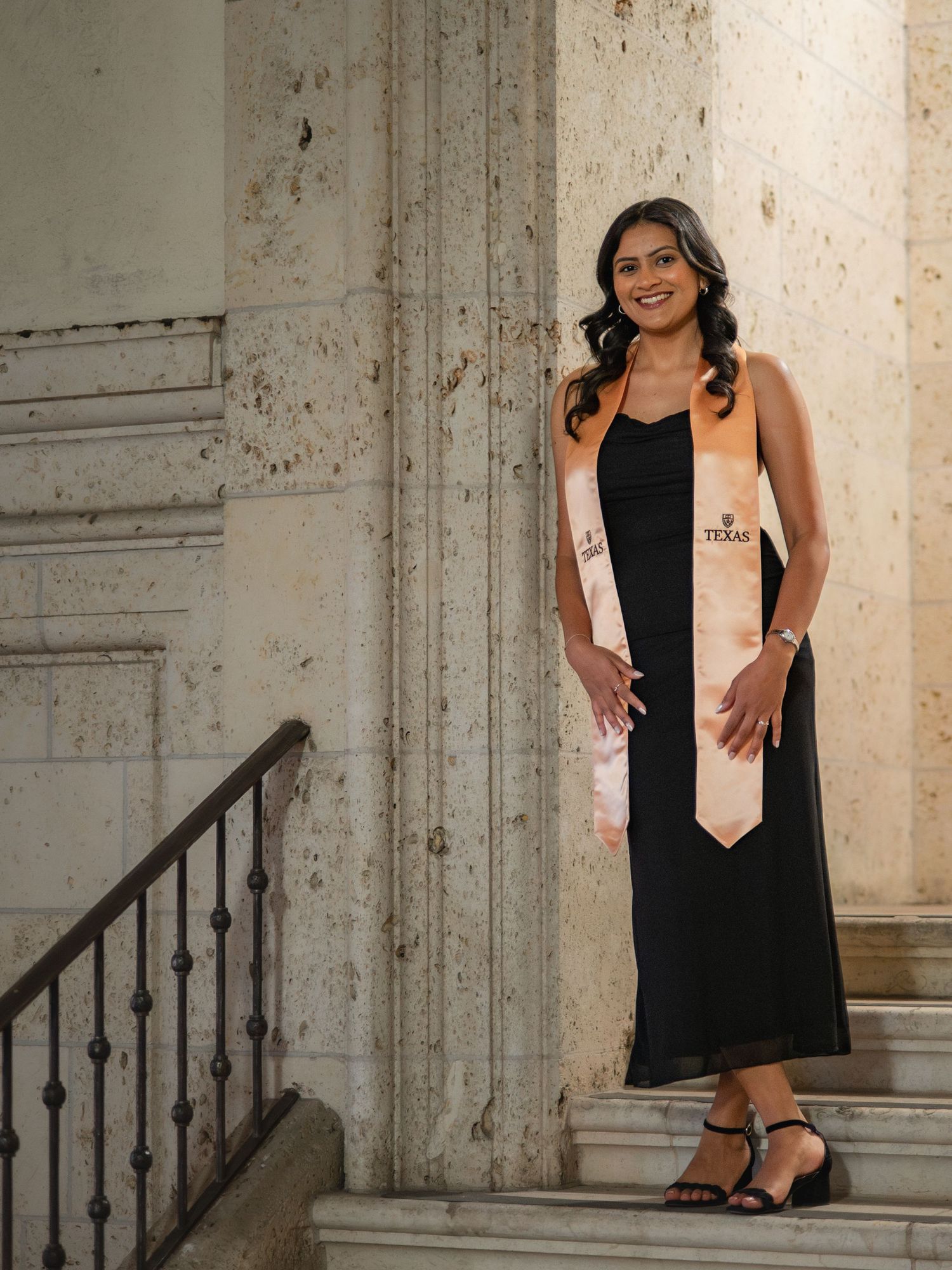 A graduate in a black dress and peach sash poses on stone steps for a series of graduation portraits.
