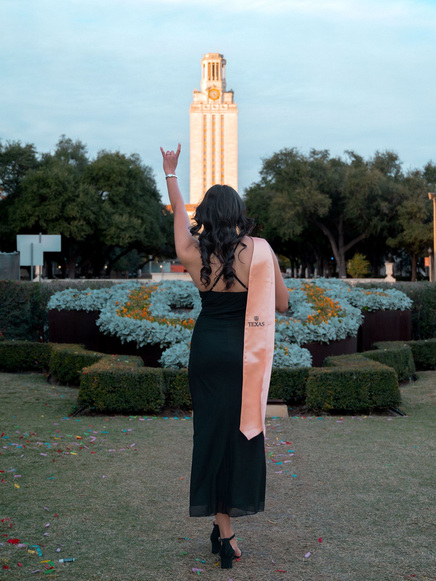 Graduate stands in front of the iconic UT Austin Tower making the Hook 'em Horns sign at sunset.
