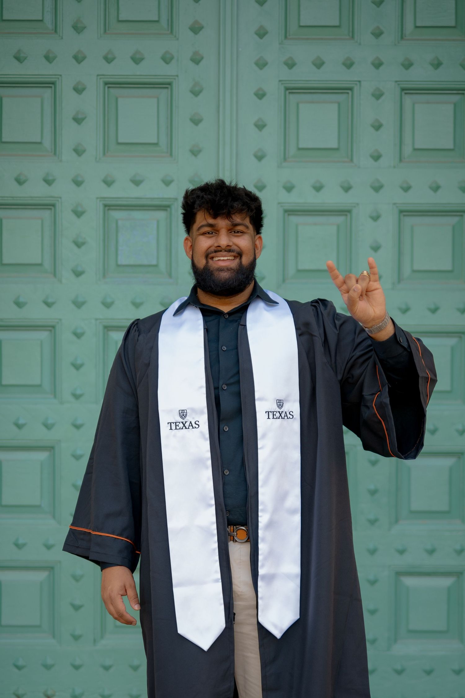 Graduate in black gown and white stole poses cheerfully by mint green doors.
