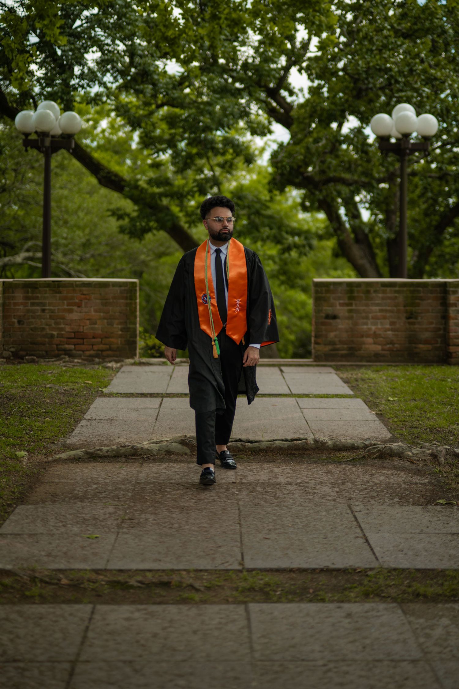 Graduate in black gown and orange stole walks along a stone path surrounded by greenery.