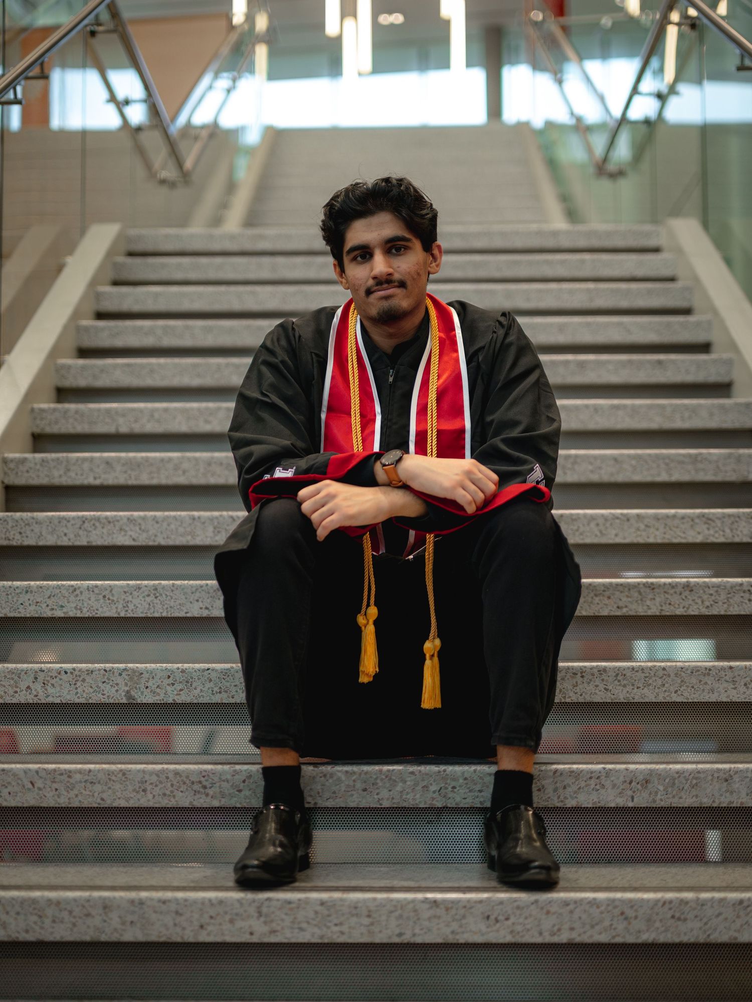 Graduate wearing black graduation attire with red stole poses on institutional steps.