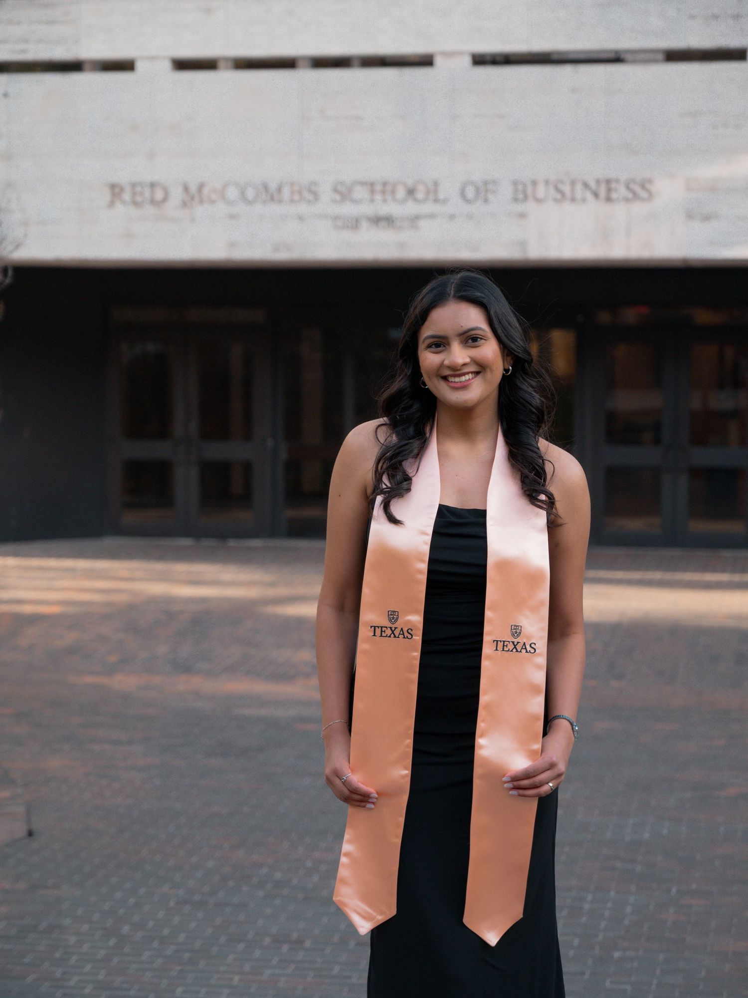 Graduate wearing black dress and peach colored stole stands in front of McCombs School of Business building.
