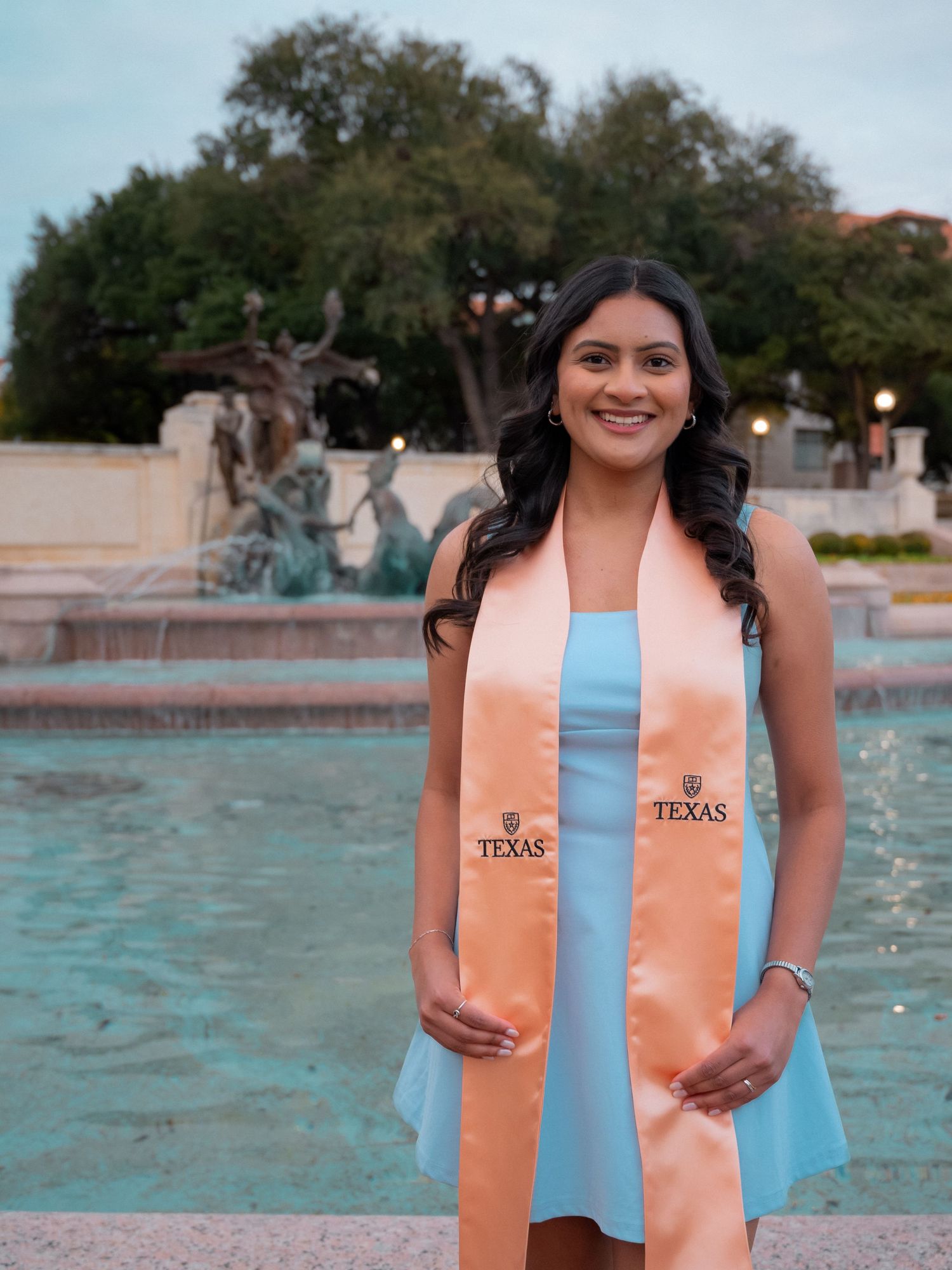 Graduate wearing orange and light blue stoles poses near a fountain during sunset celebration at University of Texas.