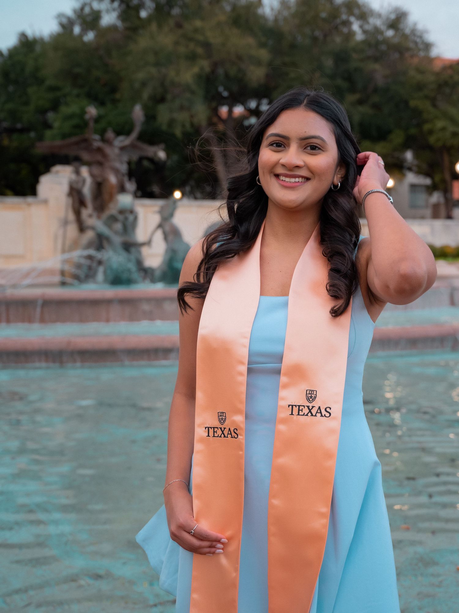 A graduate in a light blue dress and orange stole poses for photos by a fountain on a sunny day.
