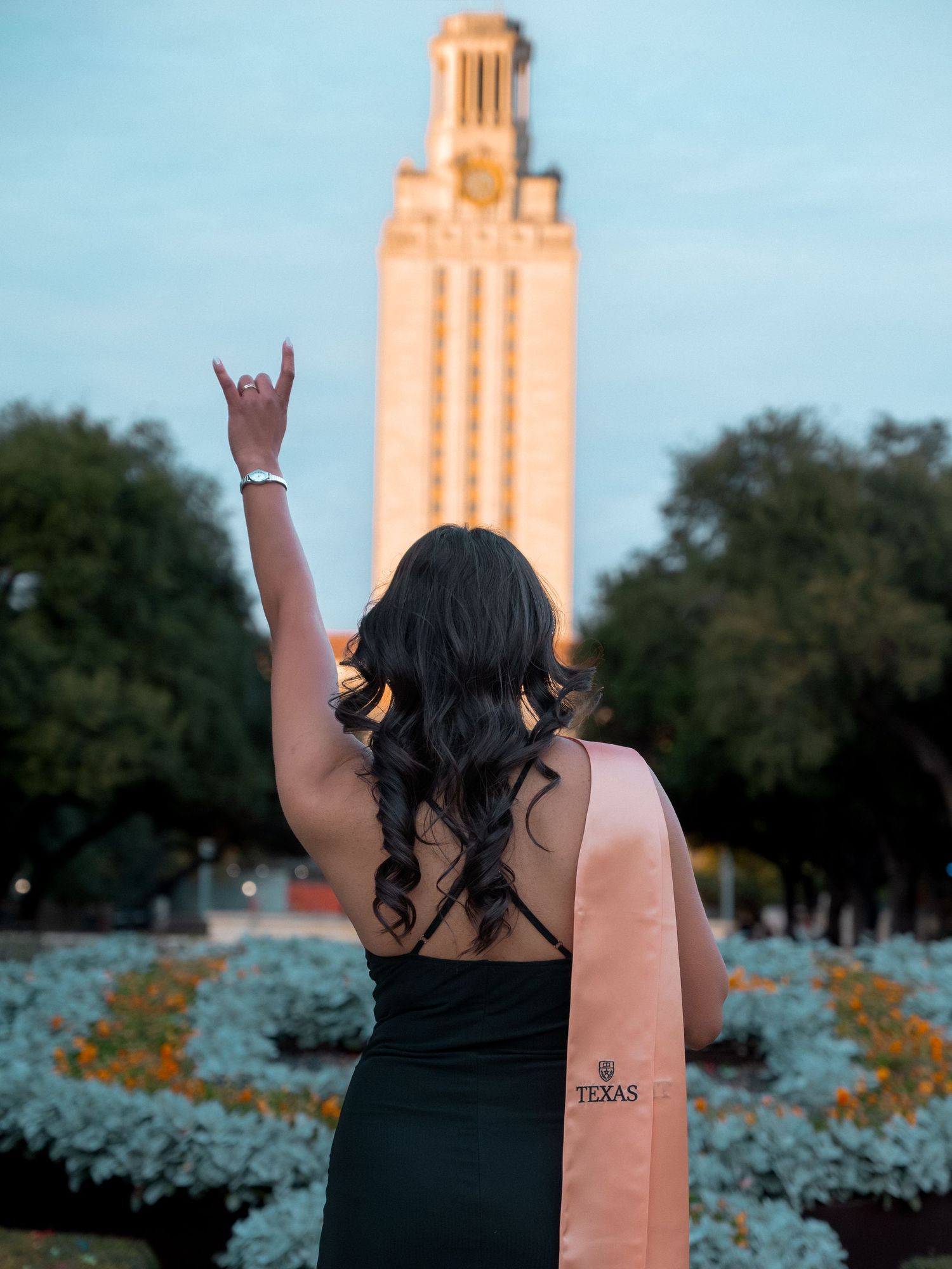 Student in black dress poses with graduation cap in front of UT Austin Tower during golden hour celebration.