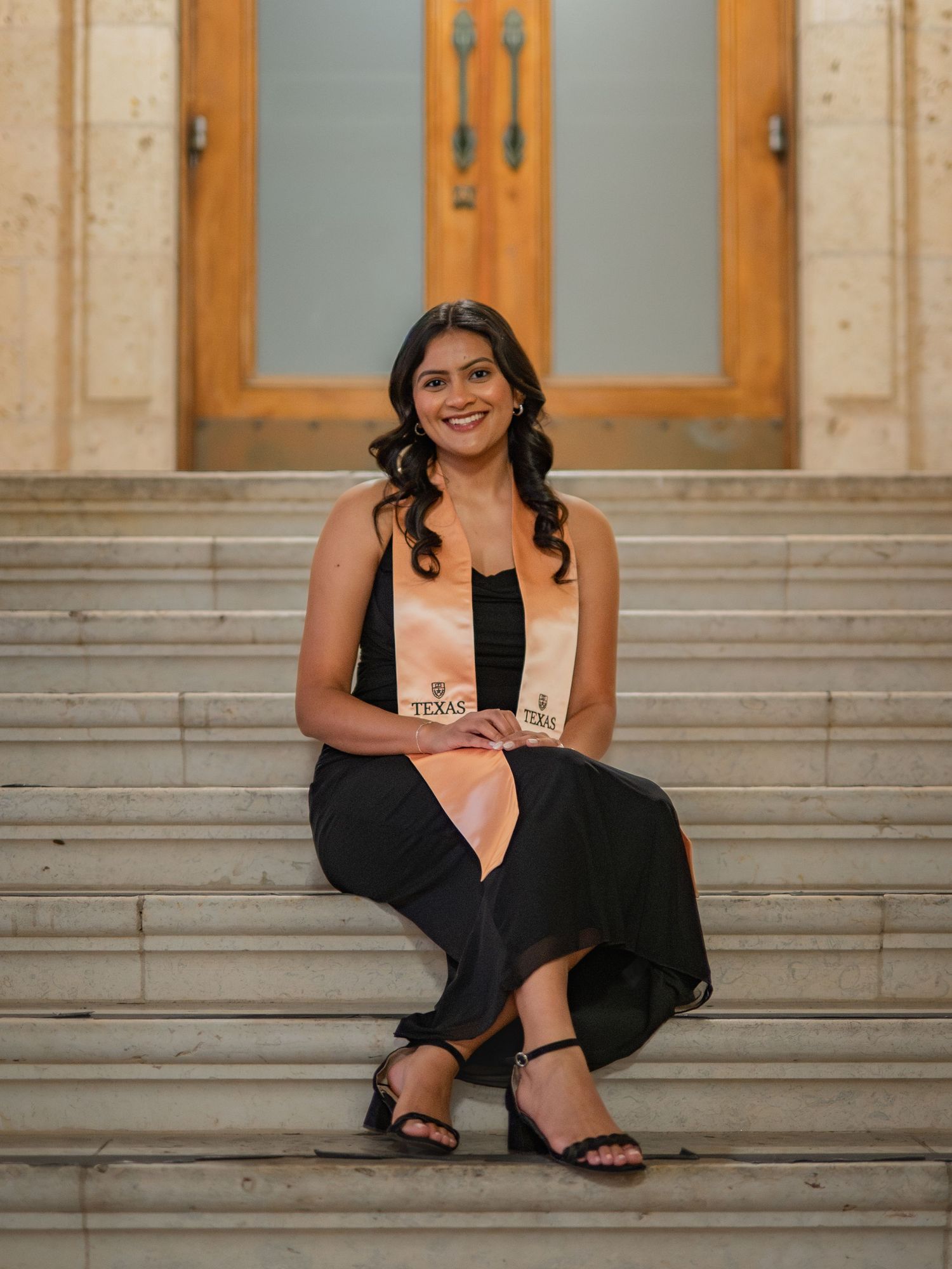 A person in a black dress sits casually on stone steps while smiling and posing for a portrait session.