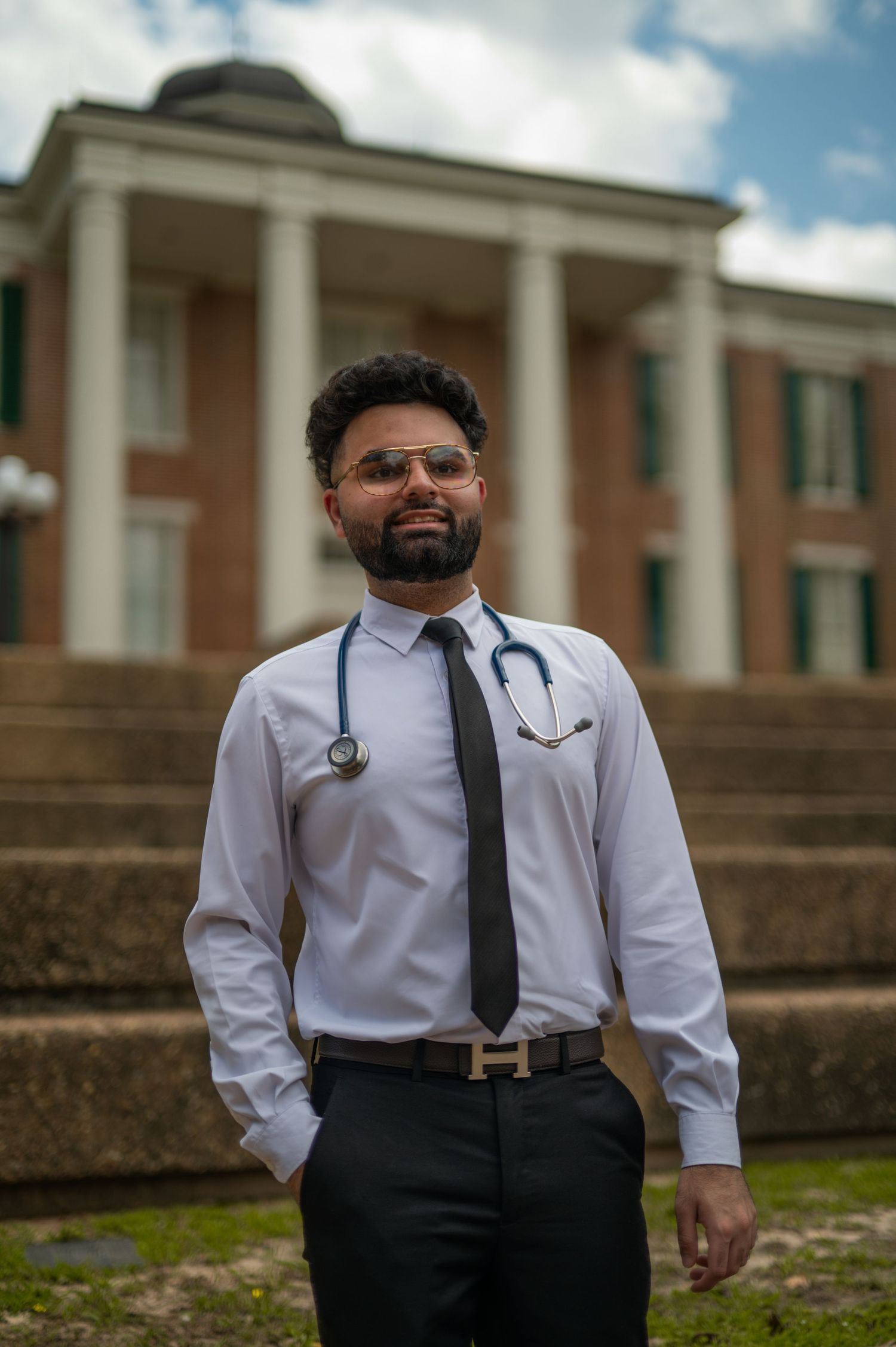 Medical professional in white coat and stethoscope poses in front of colonial-style building.