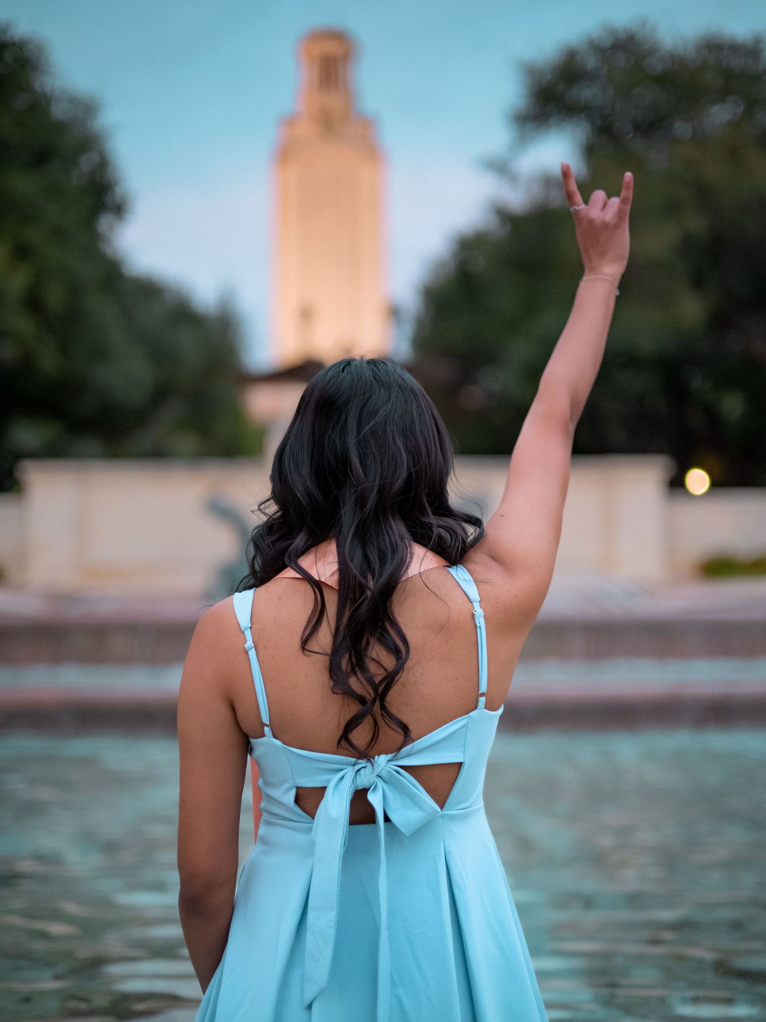 Person in light blue dress makes rock and roll hand gesture while standing by fountain with UT Austin tower in background.