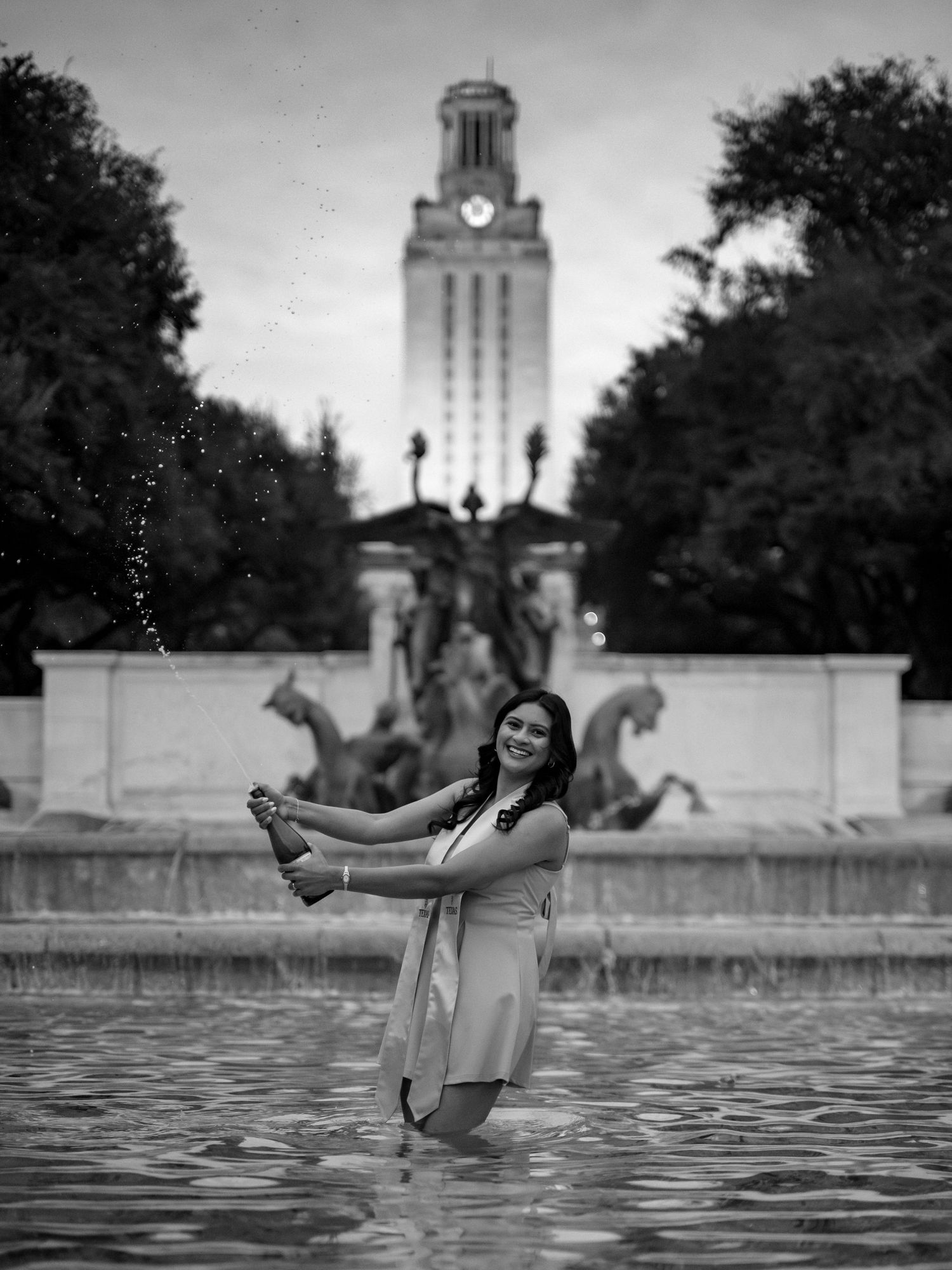 A person stands in a fountain with the iconic University of Texas Tower visible in the background at dusk.