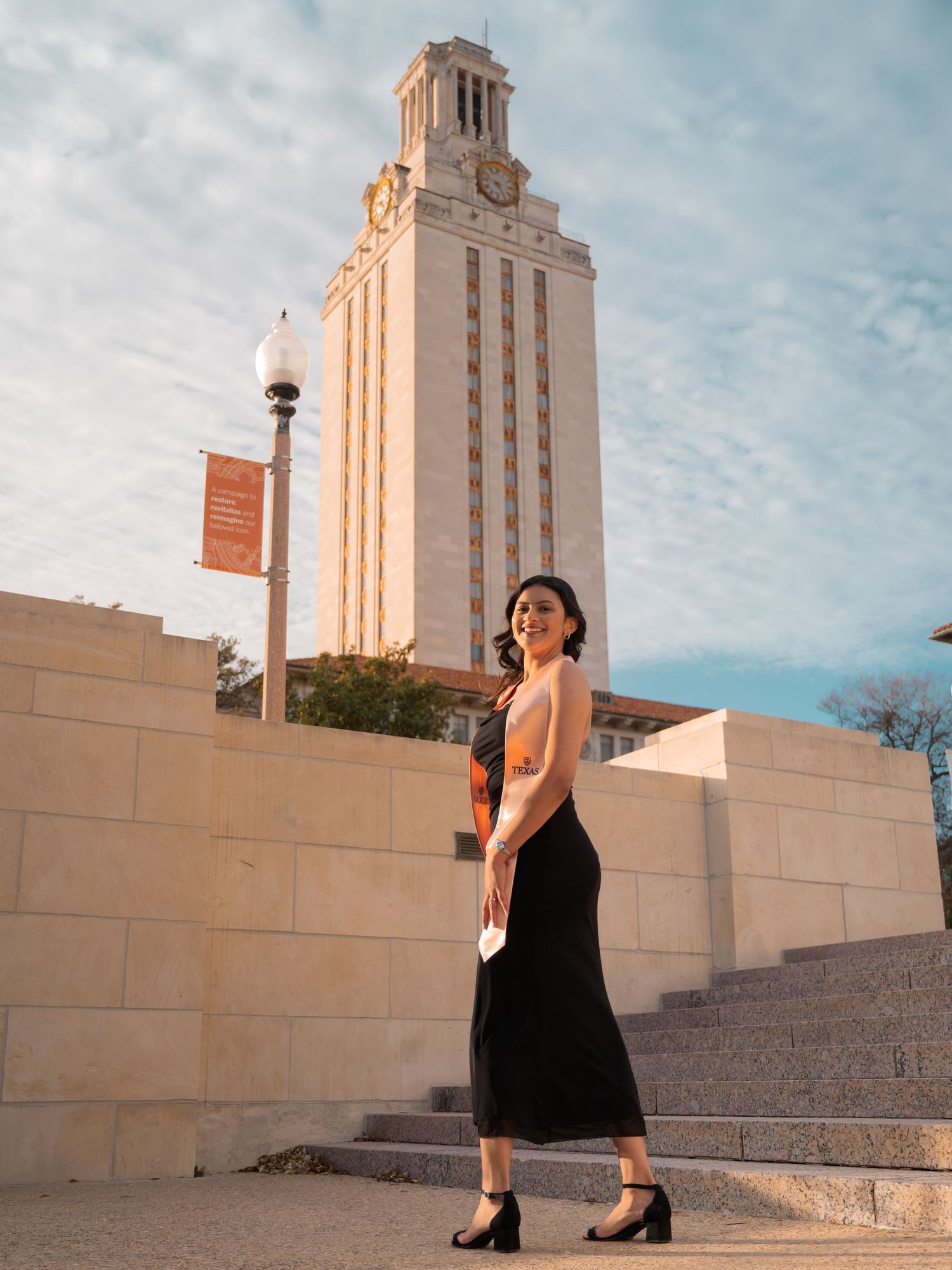 A figure in a black dress poses gracefully on concrete steps with the iconic UT Austin Tower in the background.