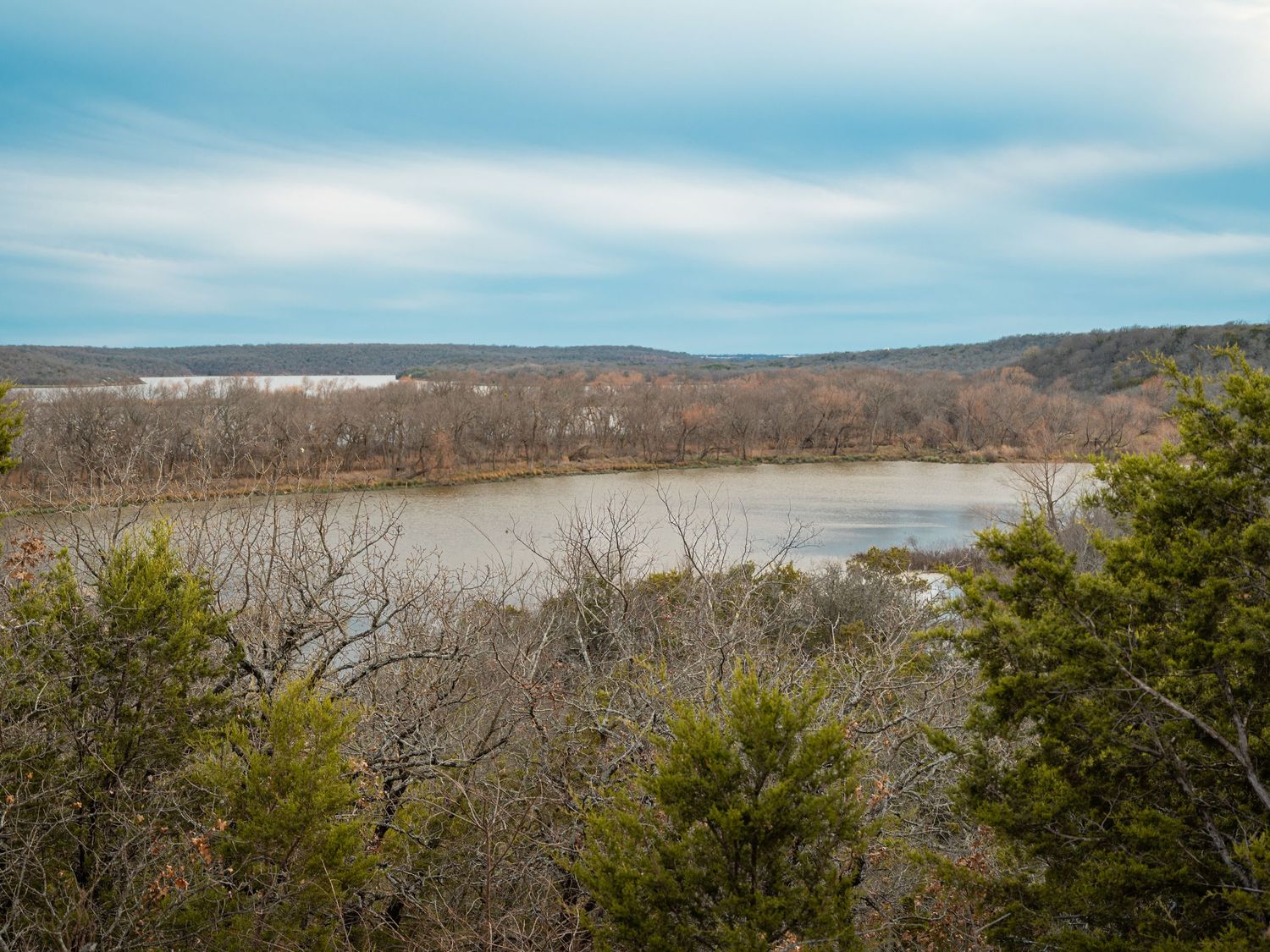 Wide panorama of a lake surrounded by hills and winter vegetation under blue skies.